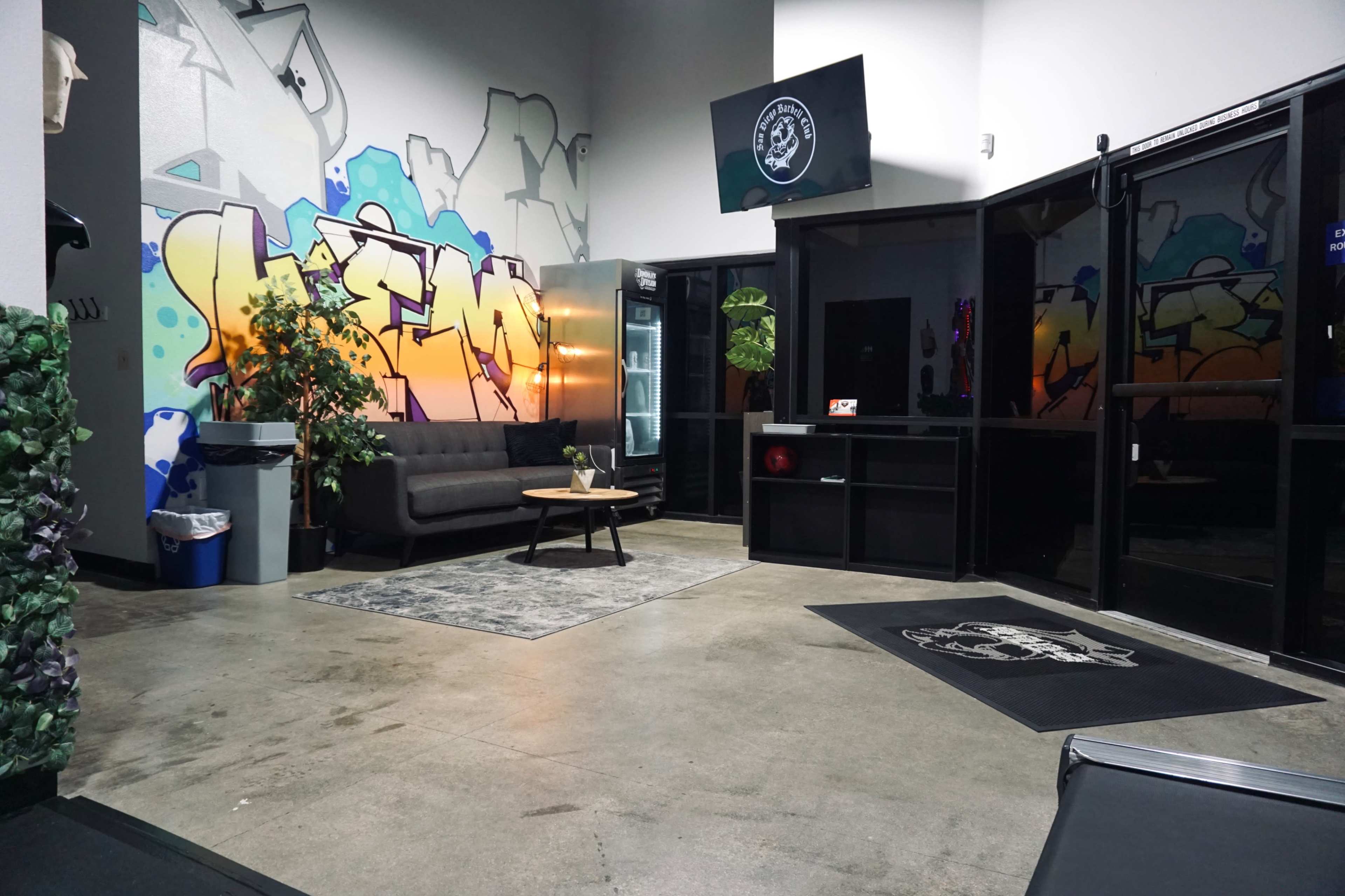 The image shows a modern lobby with a graffiti mural, a couch, a coffee table, and a visible entrance with vending machines.