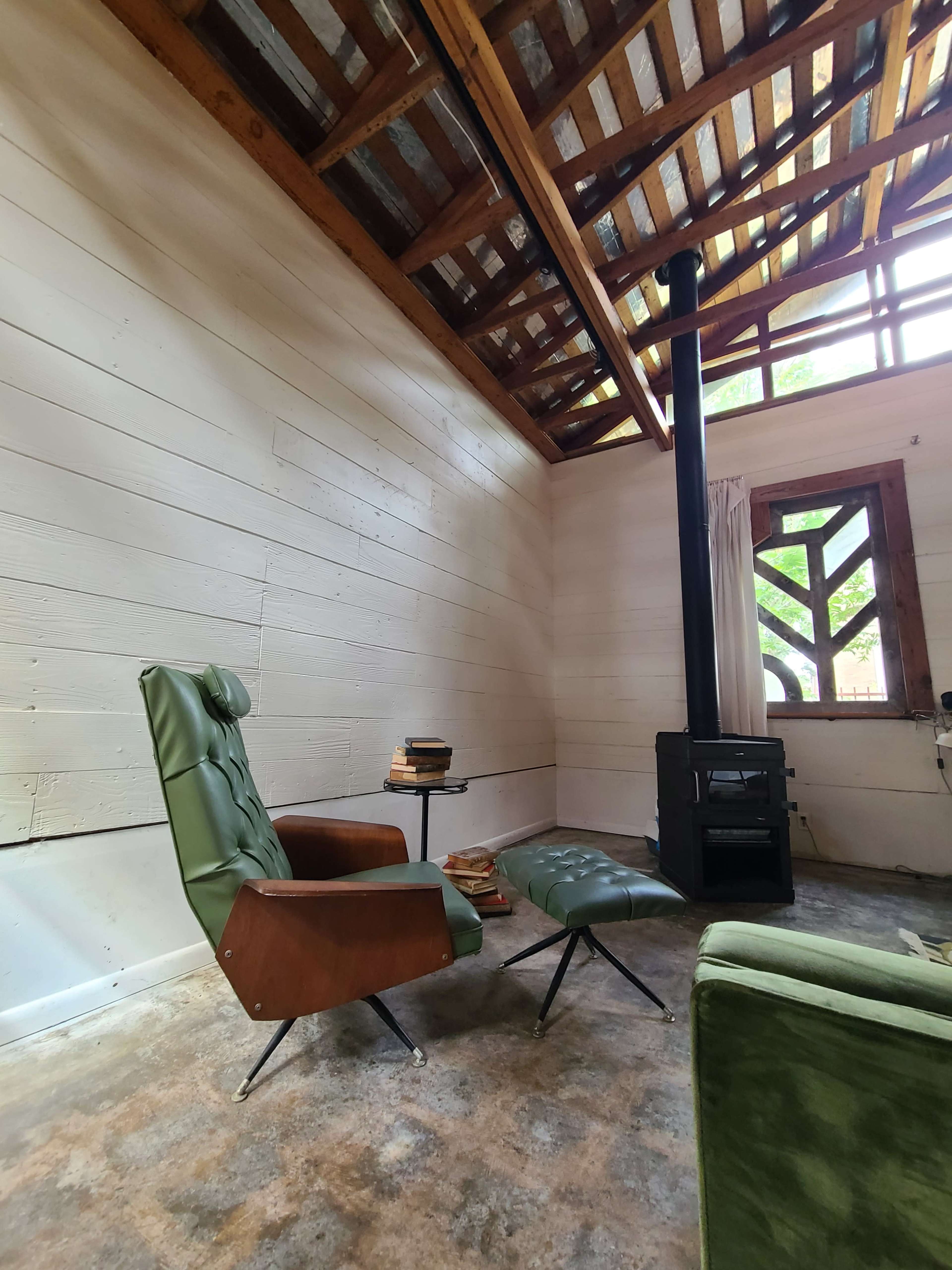 A cozy interior space featuring a green armchair and ottoman, a small side table with books, and a black wood stove, all set against a backdrop of wooden walls and a sloped roof.