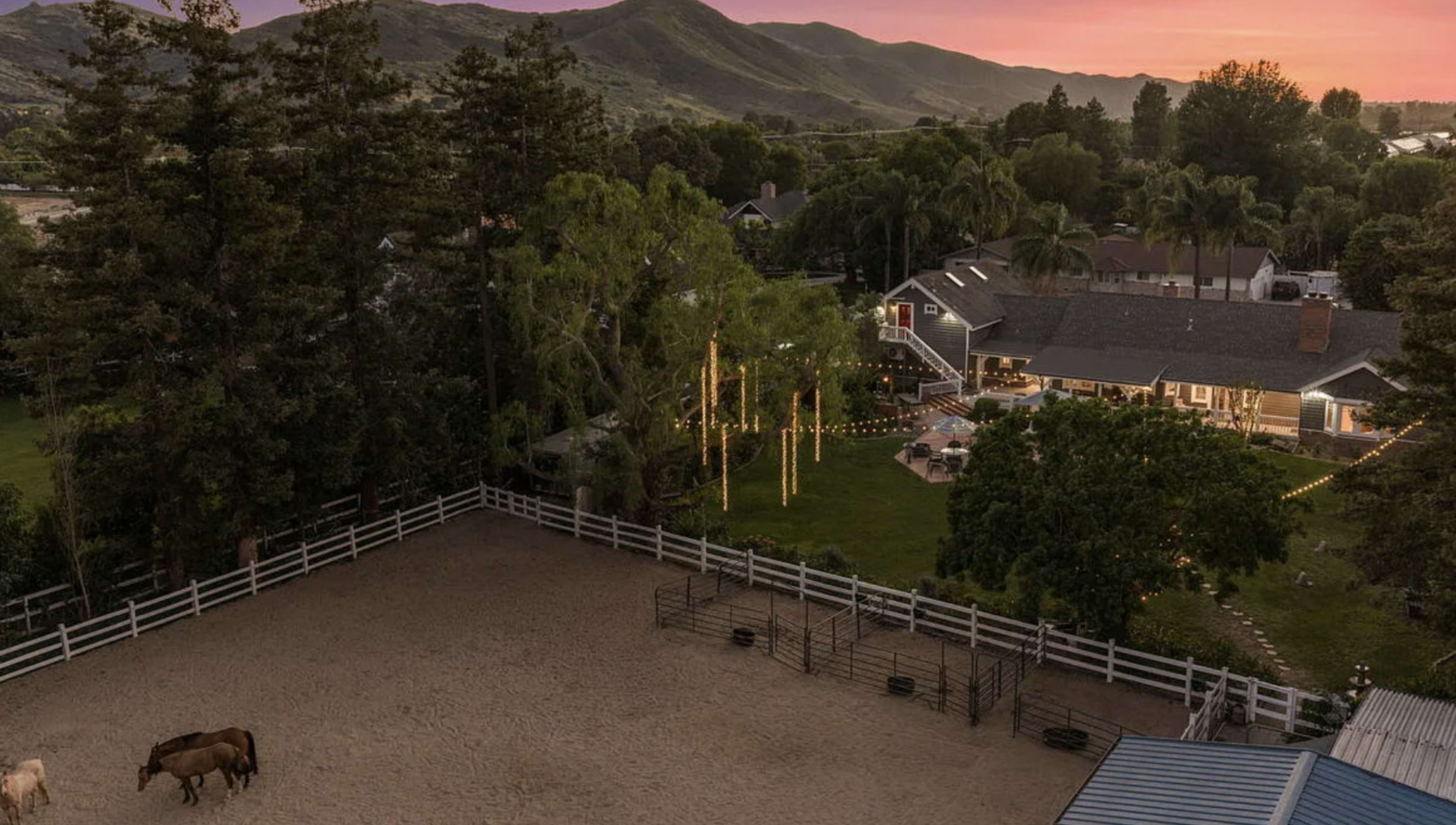 The image shows a spacious equestrian facility at dusk, featuring horses in a sandy enclosure and a house with outdoor lighting surrounded by lush trees and mountains in the background.