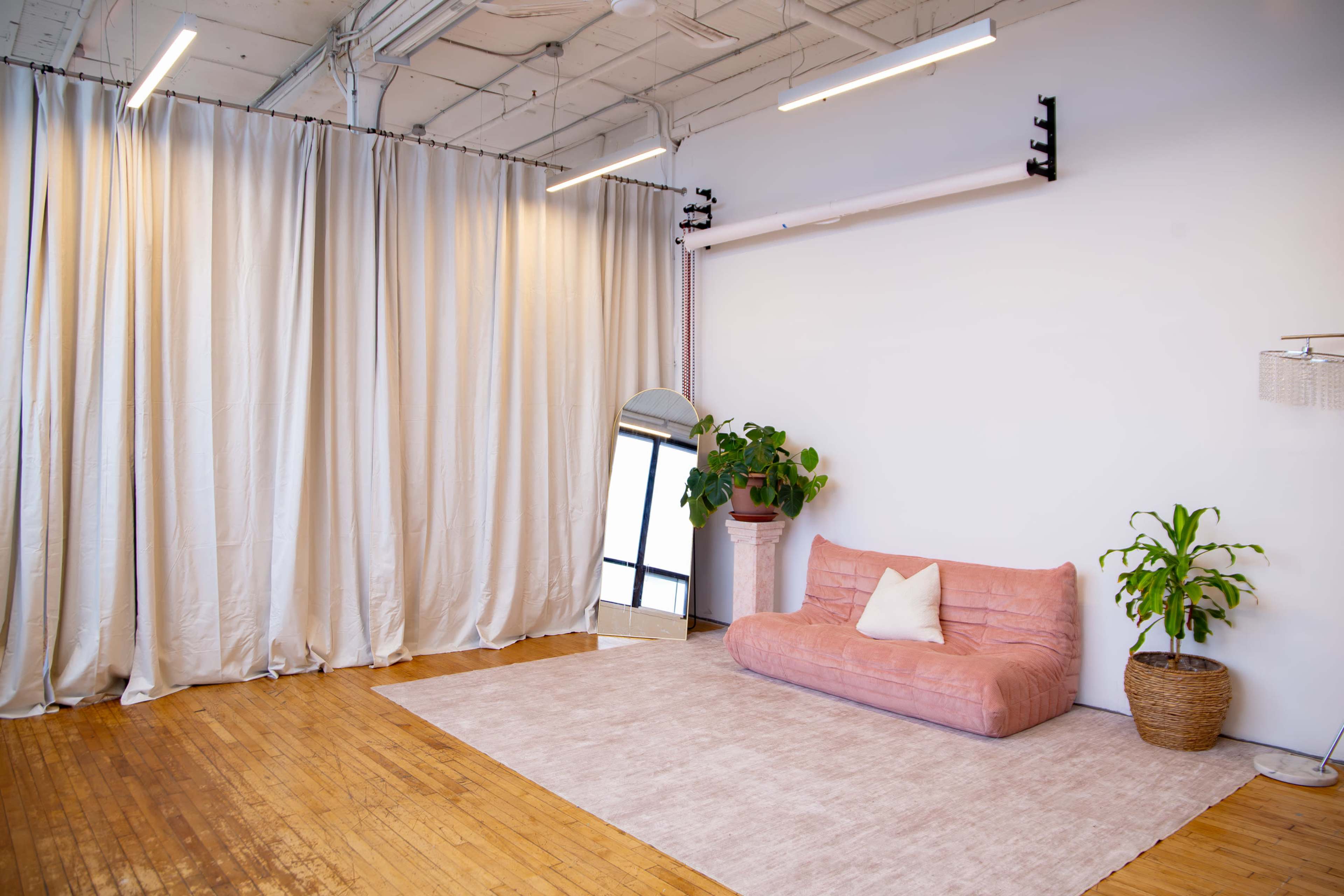 The image shows a modern interior space with a pink sofa, a mirror, a large plant, and light-colored curtains, all set on a wooden floor.
