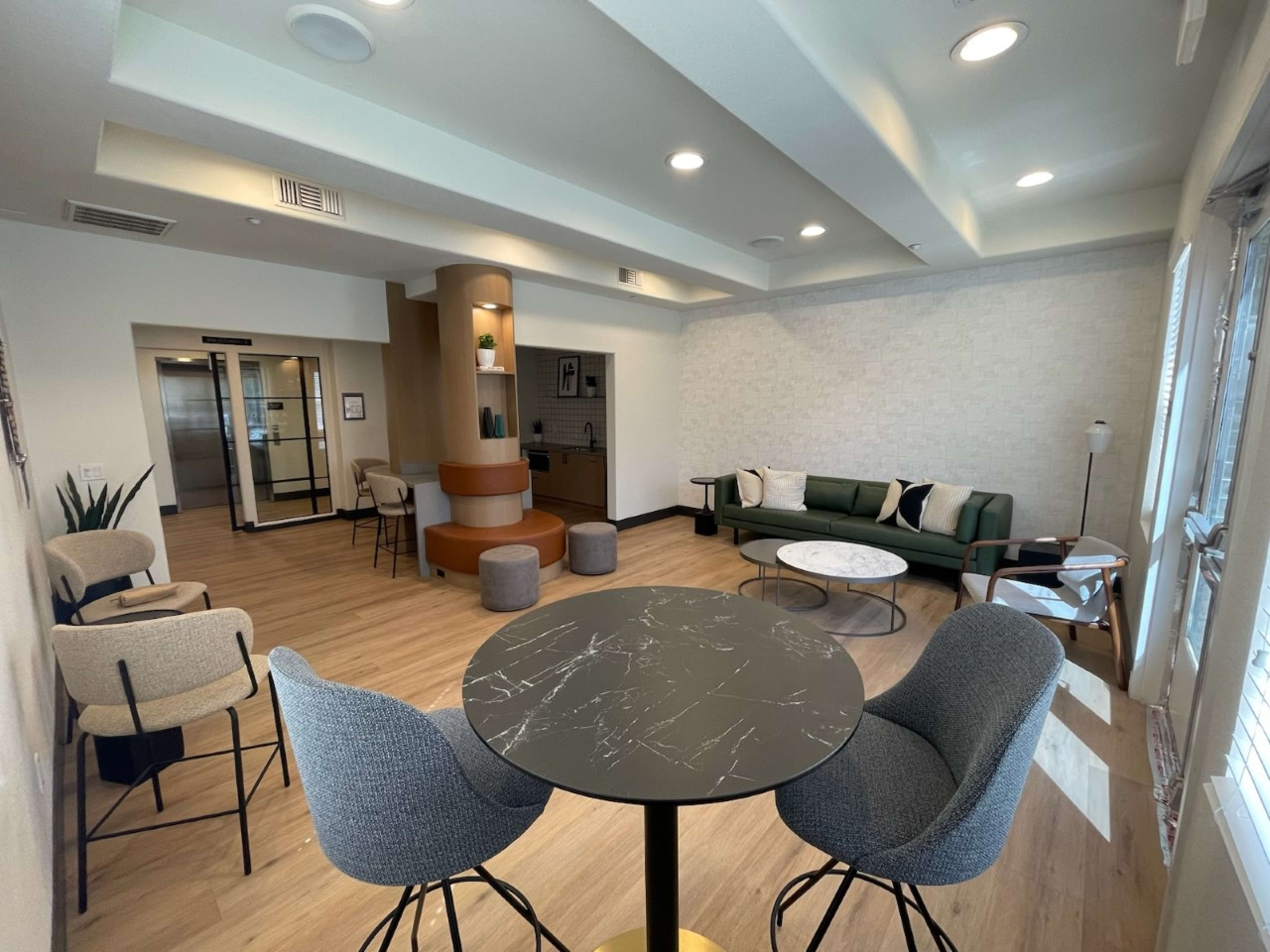 A modern lounge area featuring a circular table, two high-backed chairs, a green sofa, and a staircase leading to an upper level, with large windows allowing natural light.