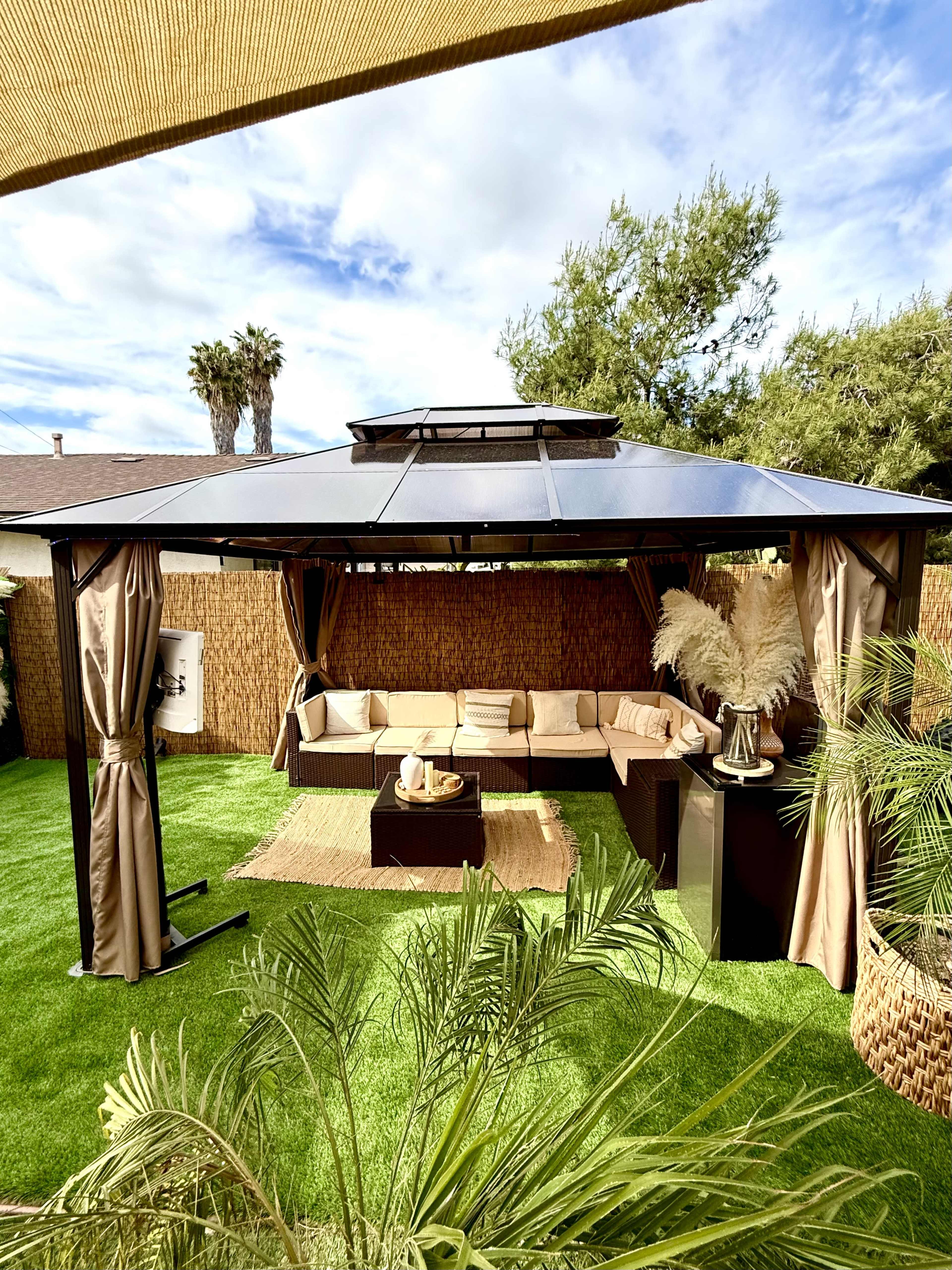 The image shows a backyard gazebo with a seating area, a low table, and decorative elements surrounded by green grass and plants.