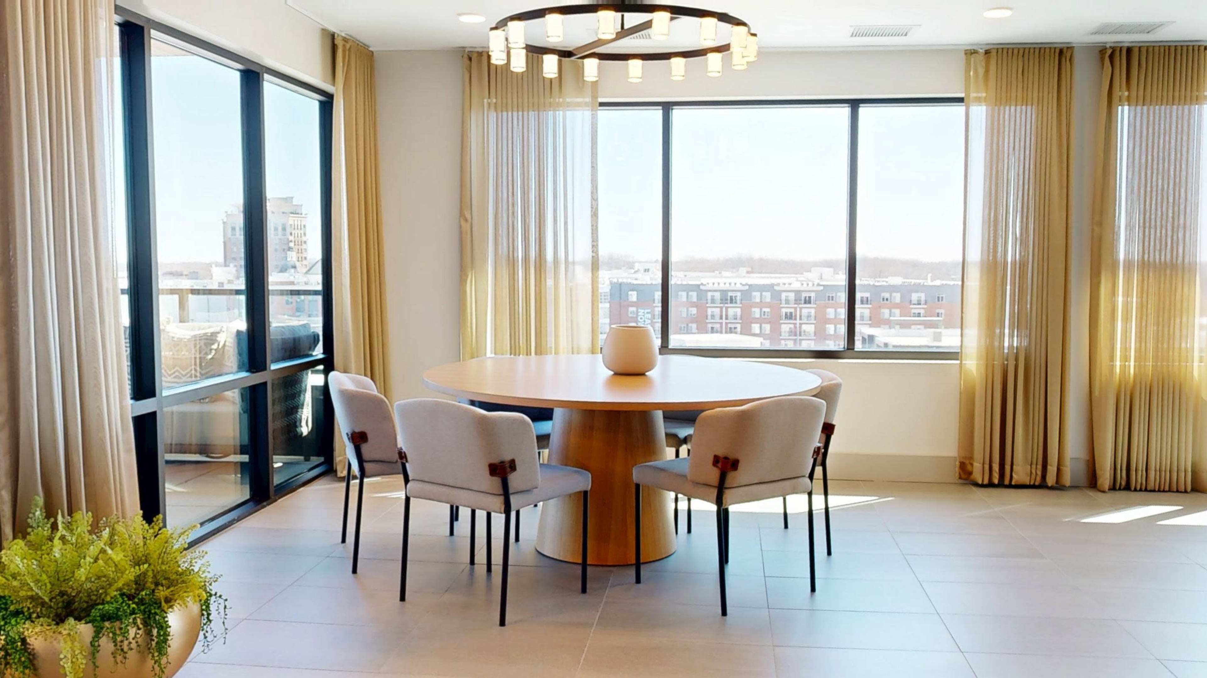 A round dining table surrounded by upholstered chairs is positioned near large windows with sheer curtains in a well-lit room.