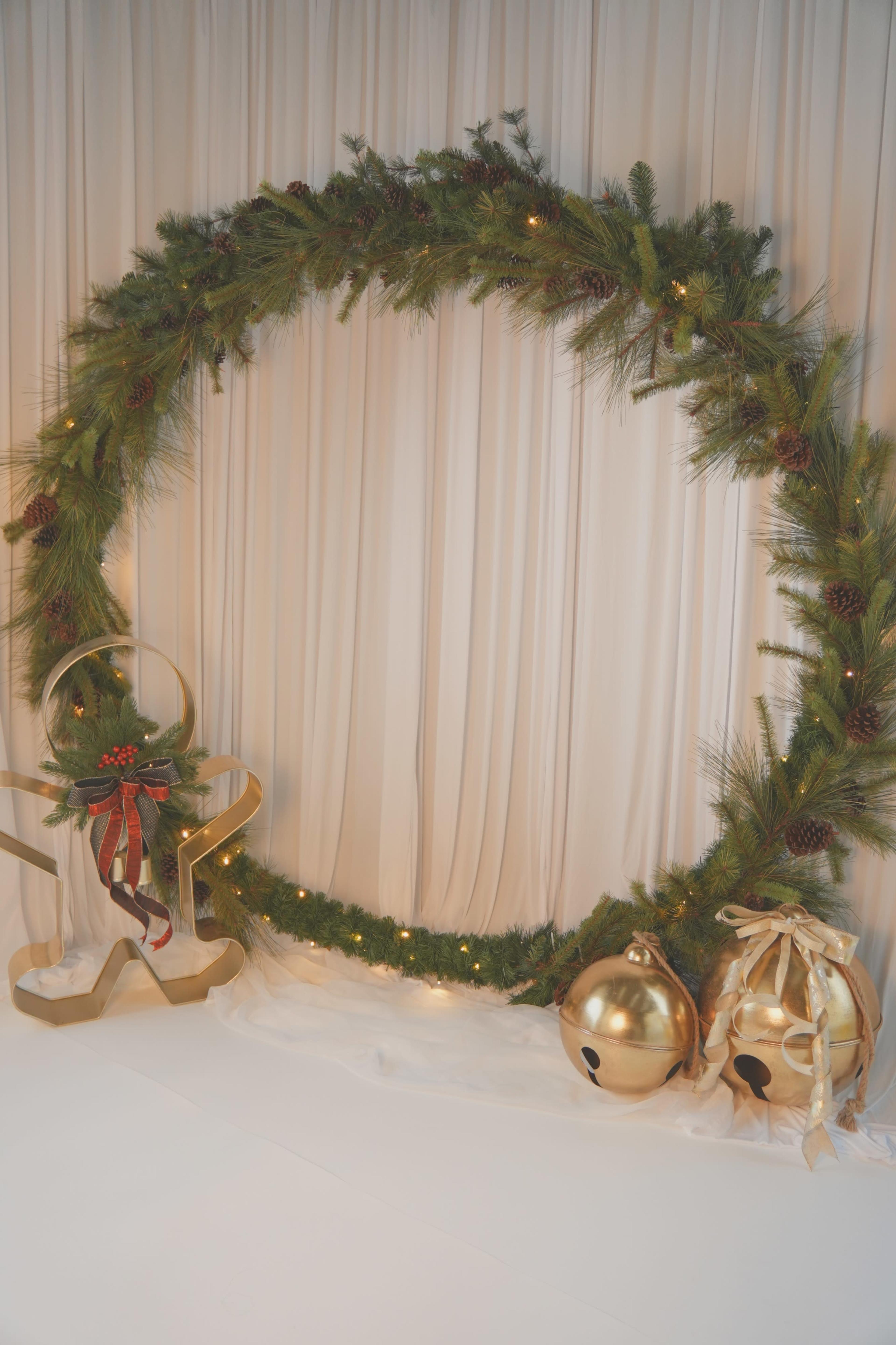 A large circular wreath made of pine branches and decorated with pinecones and lights stands against a curtain backdrop, accompanied by a pair of gold jingle bells and a decorative sleigh.