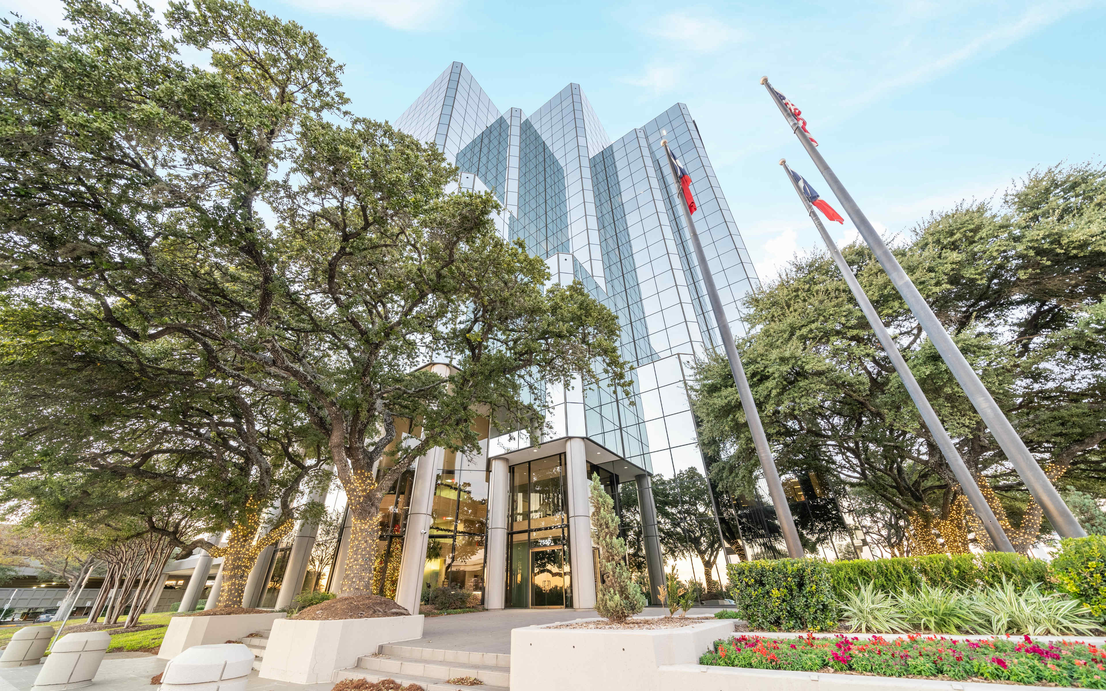 A modern glass office building stands surrounded by trees and flagpoles, with landscaped greenery in the foreground.