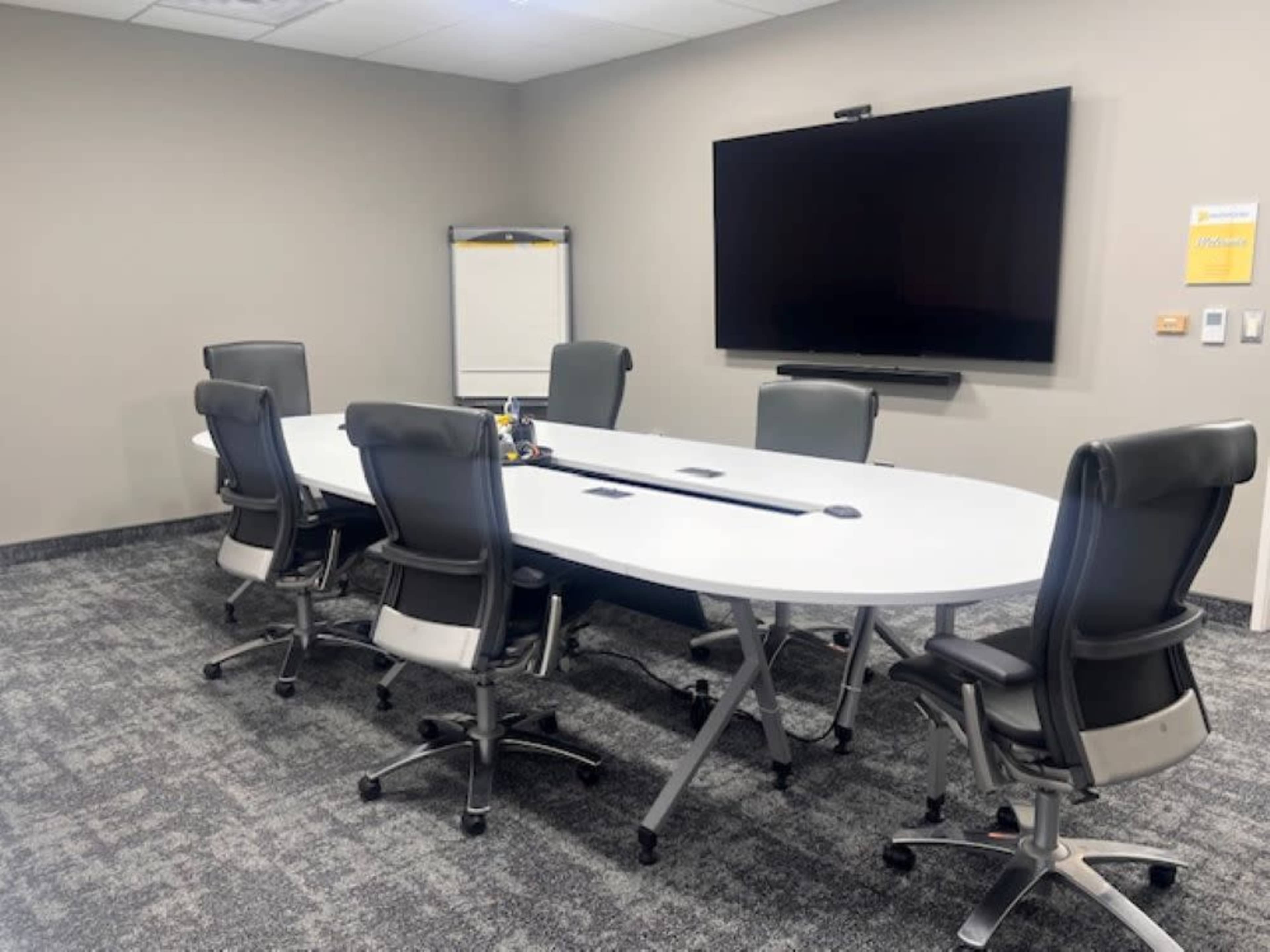 The image shows a conference room with a large oval table surrounded by six ergonomic chairs, a wall-mounted screen, and a flip chart stand.