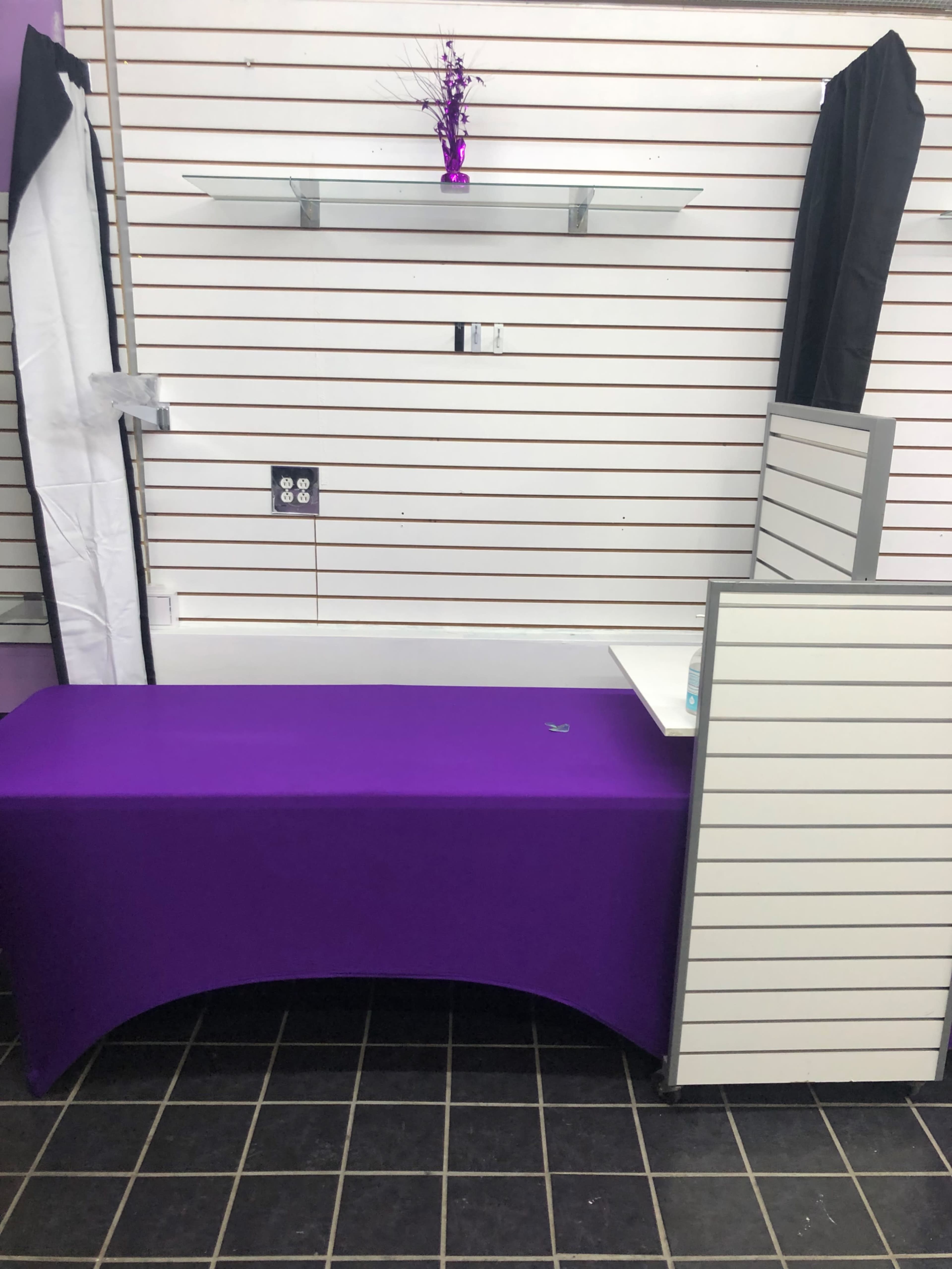 A purple table with a decorative item sits in front of a wall featuring slatboard and shelves.