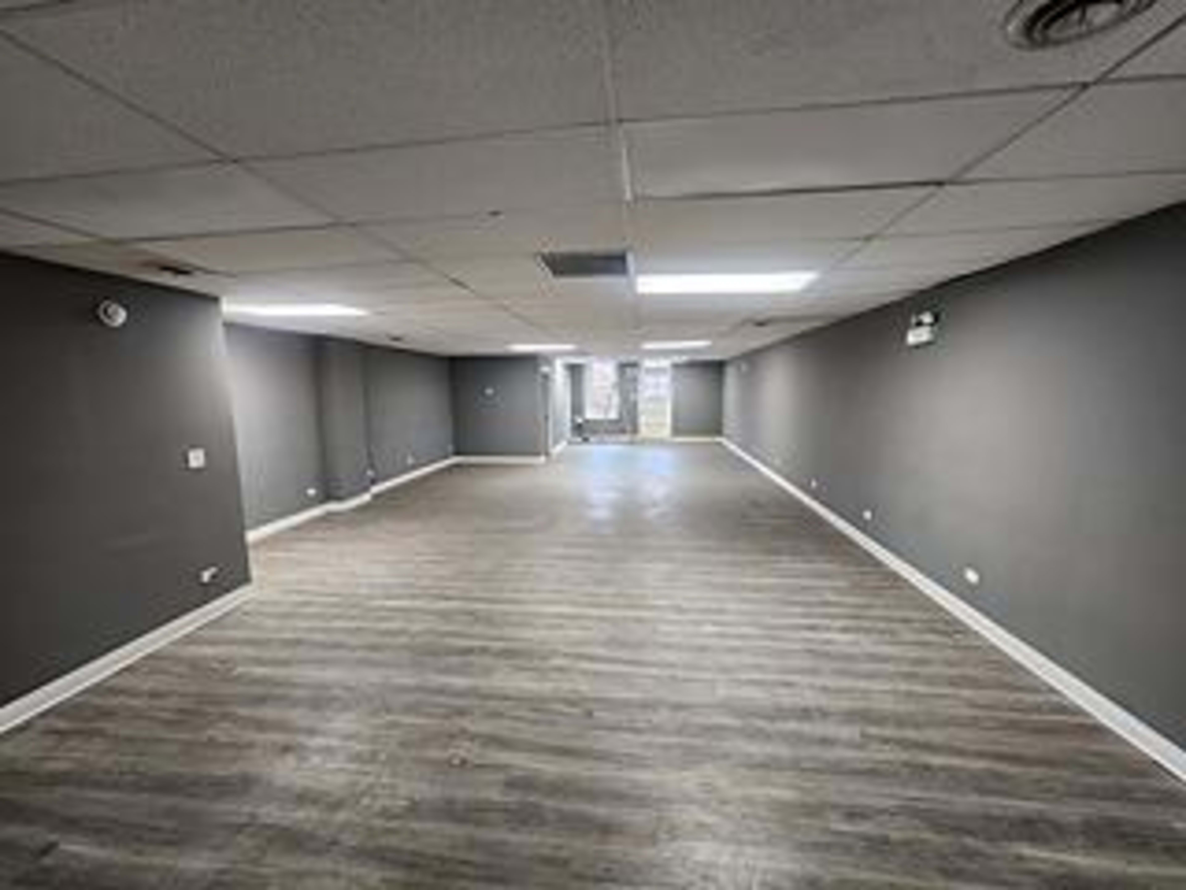 The image shows a spacious, empty interior room with gray walls and wooden flooring, illuminated by overhead lights.