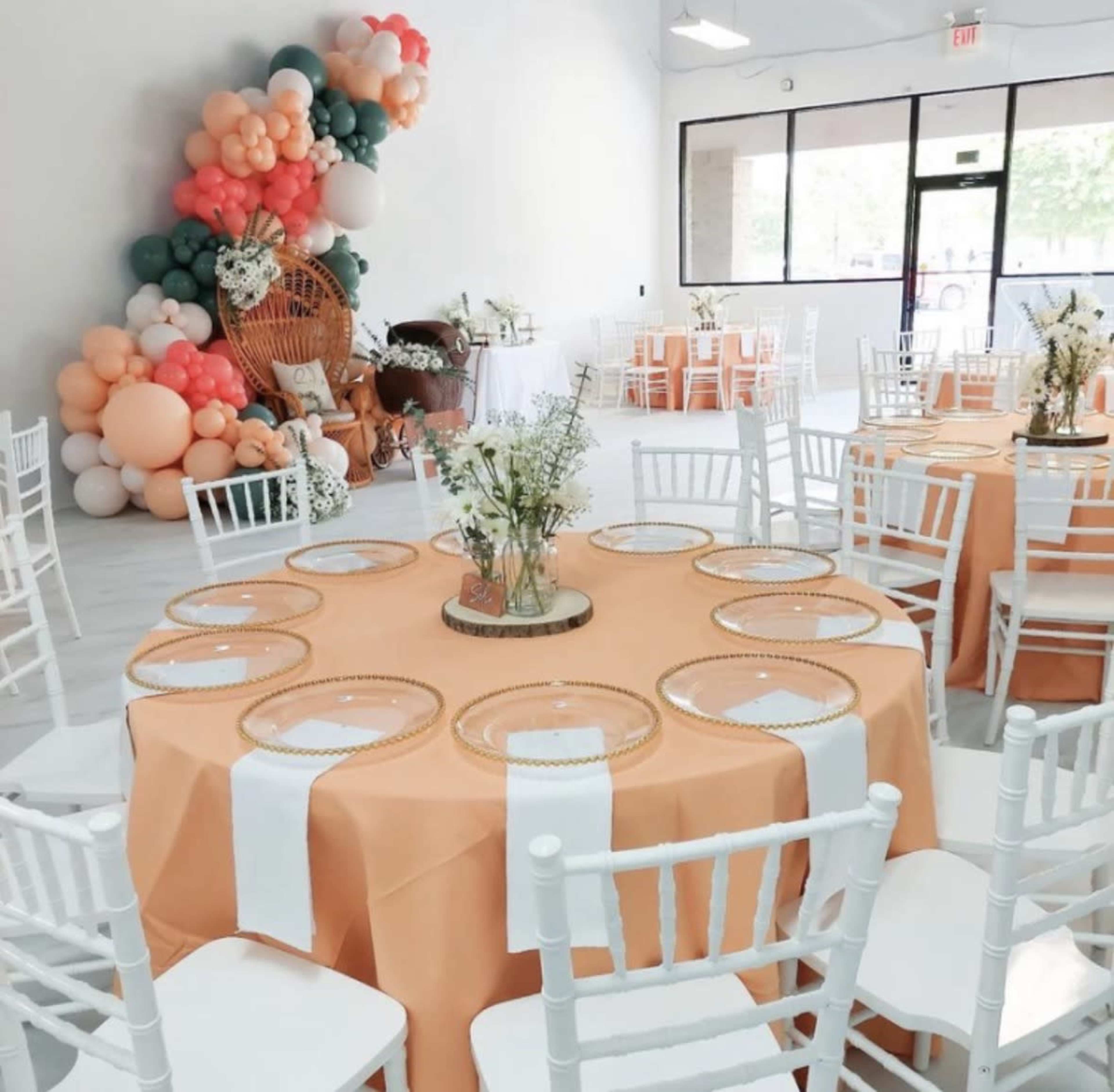 The image shows a decorated indoor event space featuring orange tablecloths, white chairs, floral centerpieces, and a colorful balloon display in the corner.
