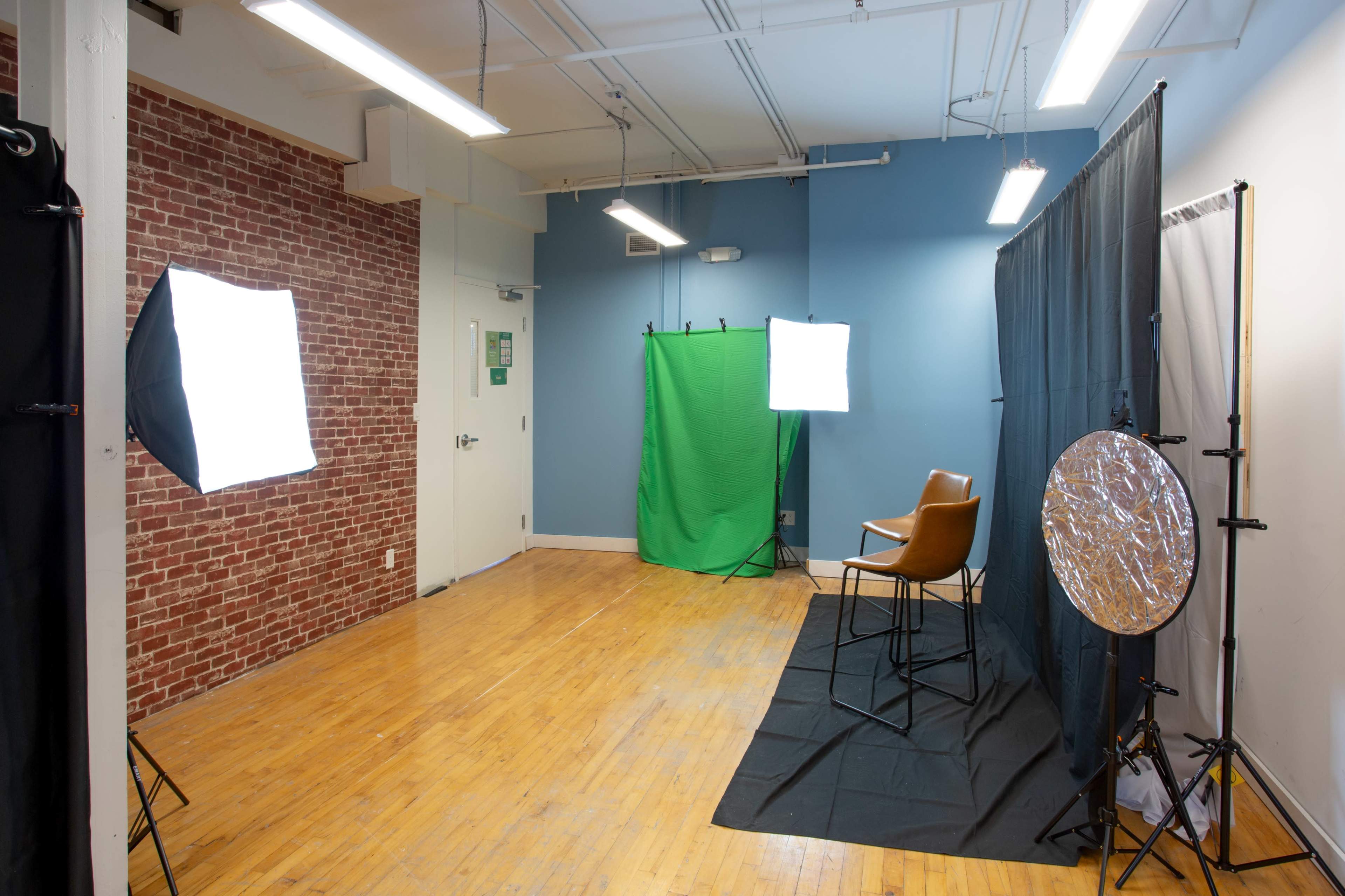 A photography studio setup featuring two light sources, a green backdrop, a folding chair, and a reflector on the floor, with brick and blue walls.
