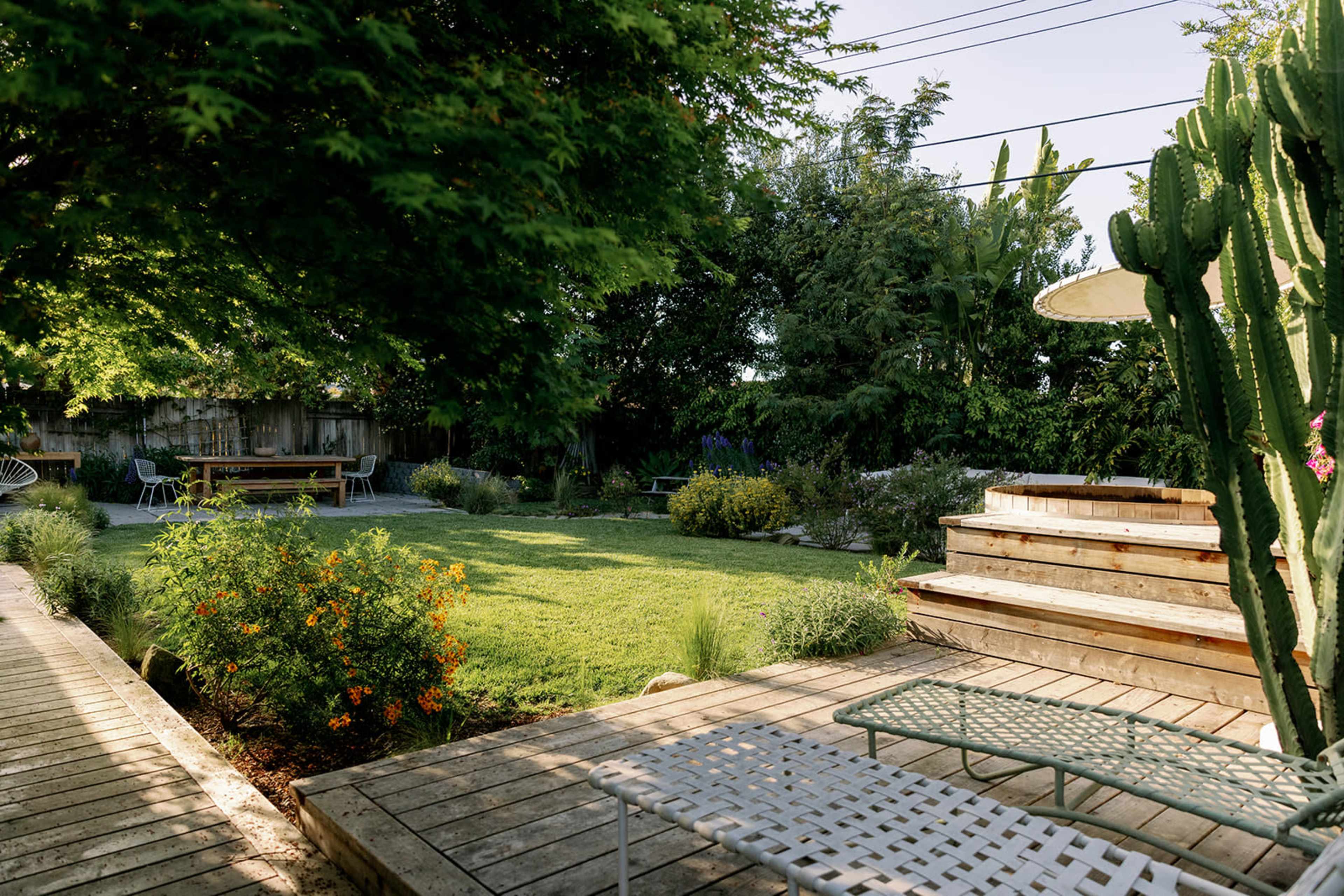 The image shows a landscaped backyard featuring a wooden deck, lounge chairs, and a grassy area surrounded by various plants and trees.