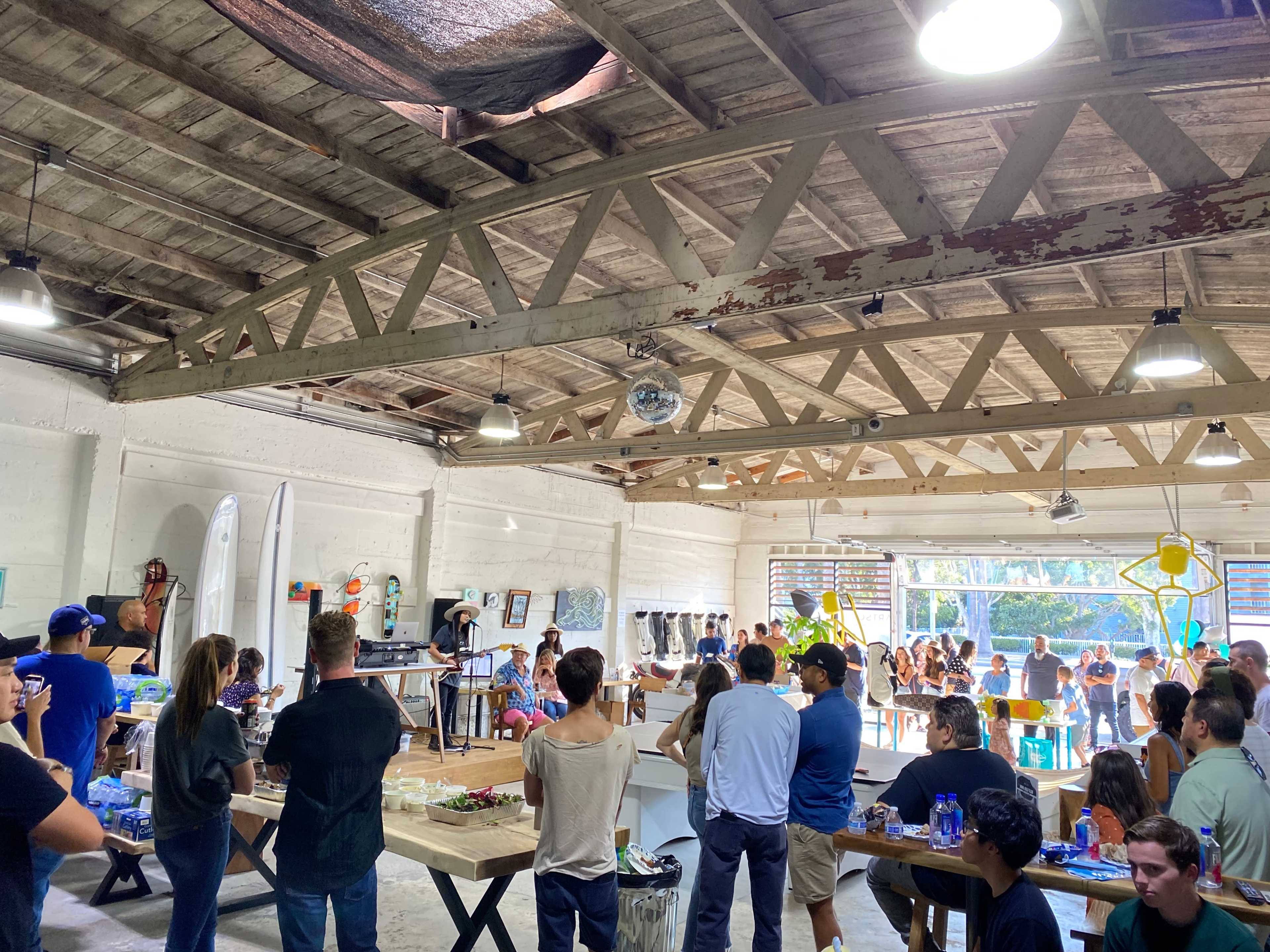 A large group of people gathers in a spacious, well-lit indoor area with surfboards on display and tables set up with food and drinks.