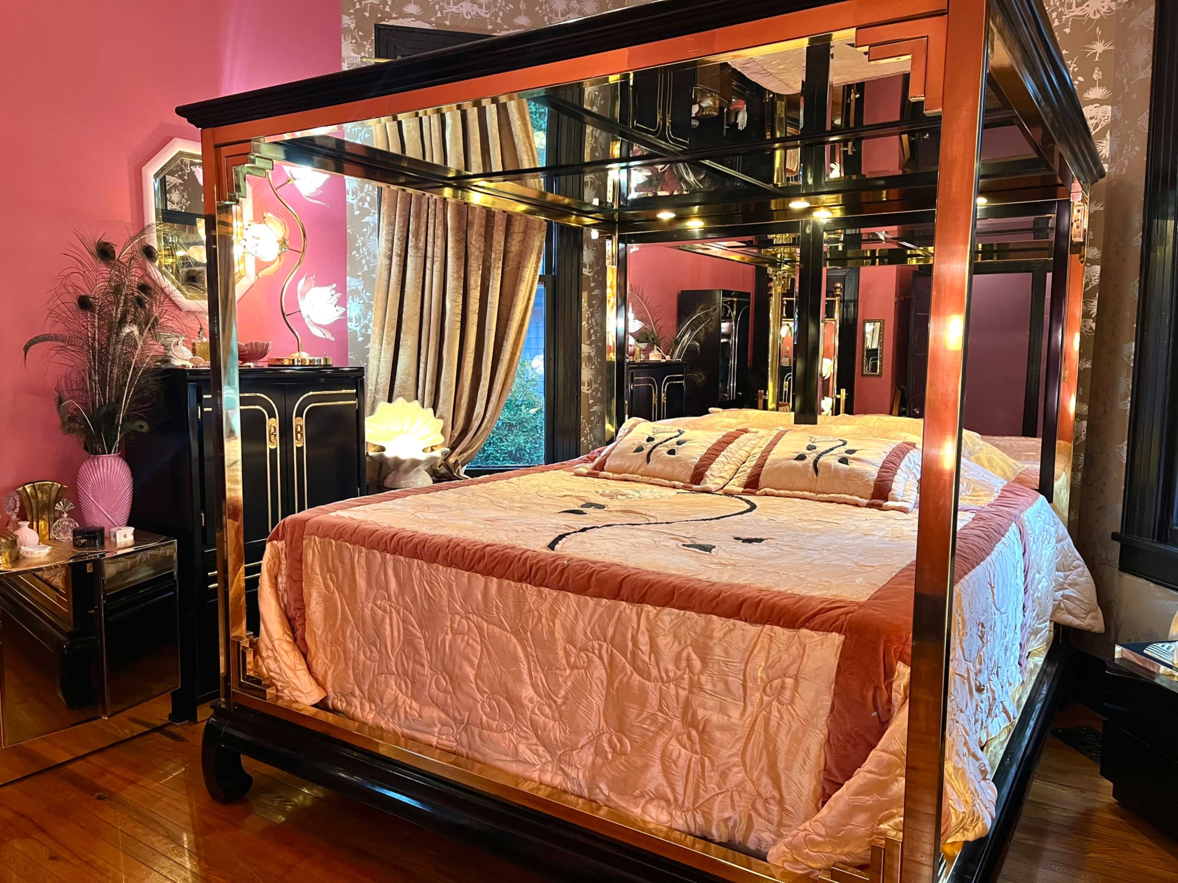 The image shows a stylish bedroom featuring a four-poster bed with a mirrored frame, ornate bedding, and elegant decor against a pink wall.