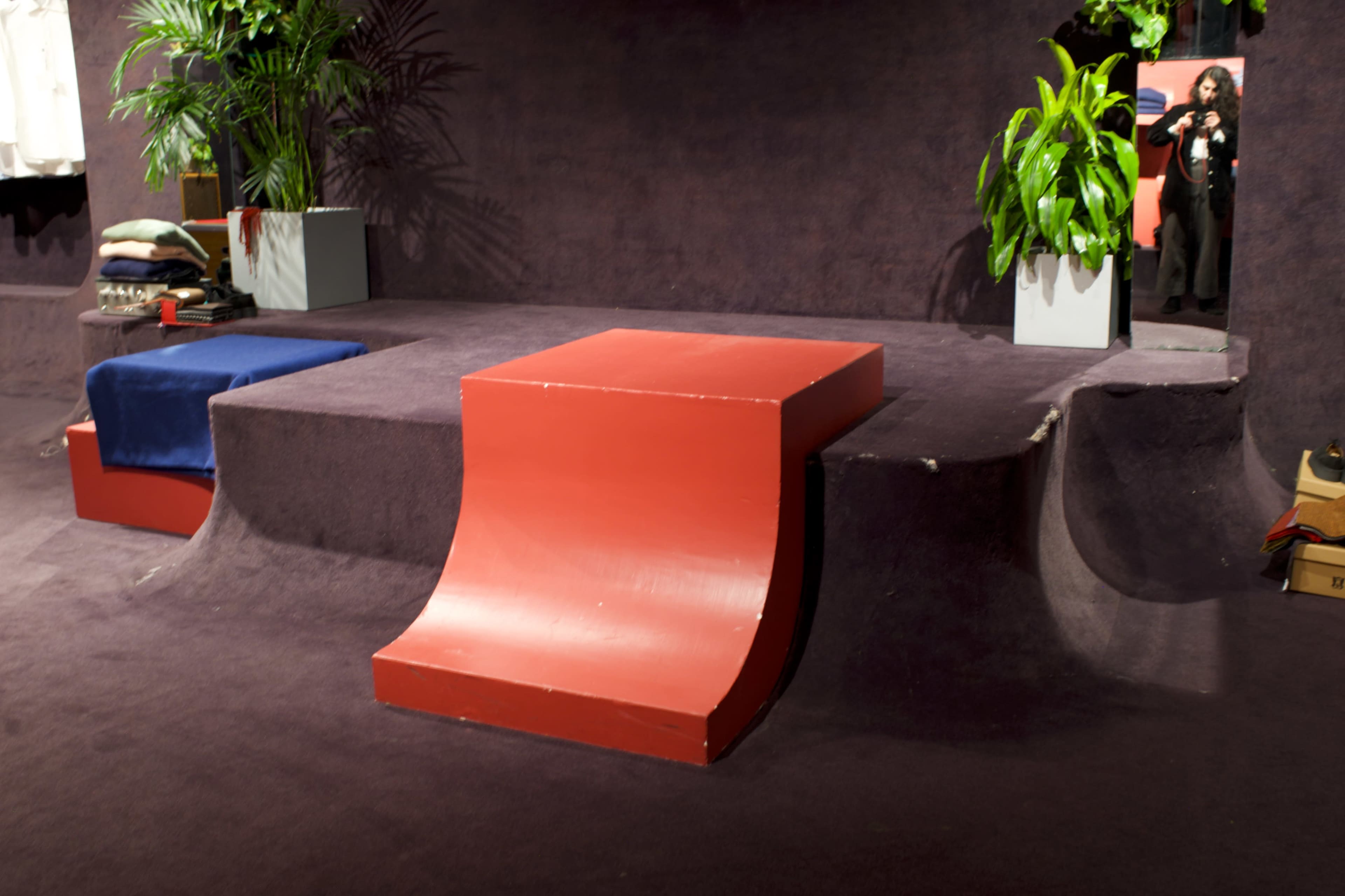 The image shows a modern space featuring a red ramp-like structure surrounded by plush carpets, with plants and clothing on display in the background.