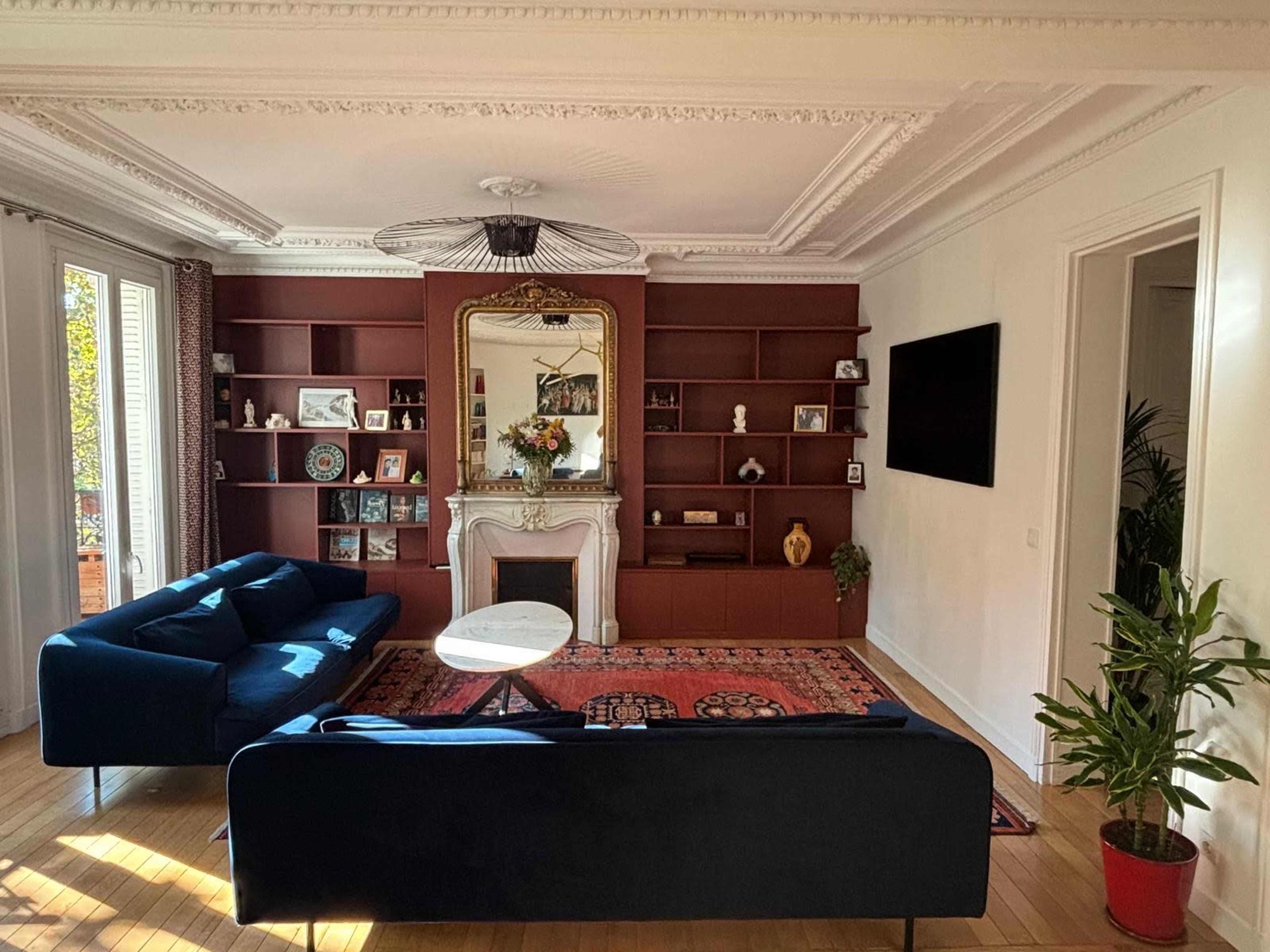 A living room features a blue sofa, a round coffee table, a decorative fireplace with a mirror above it, and bookshelves filled with various objects.