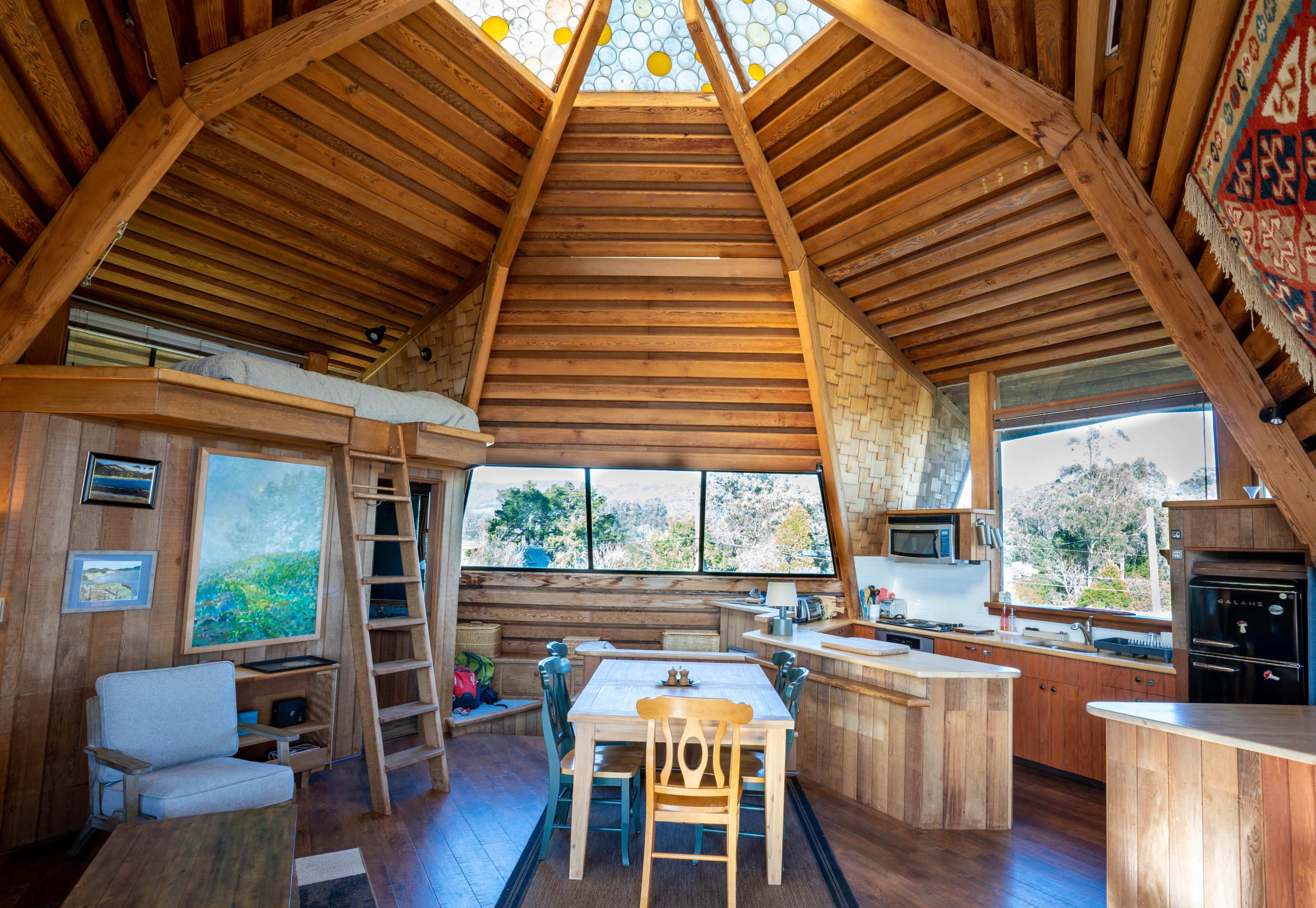 The interior of a wooden, triangular cabin features a kitchen, dining area, and a lofted sleeping space, with large windows allowing natural light to flood the room.