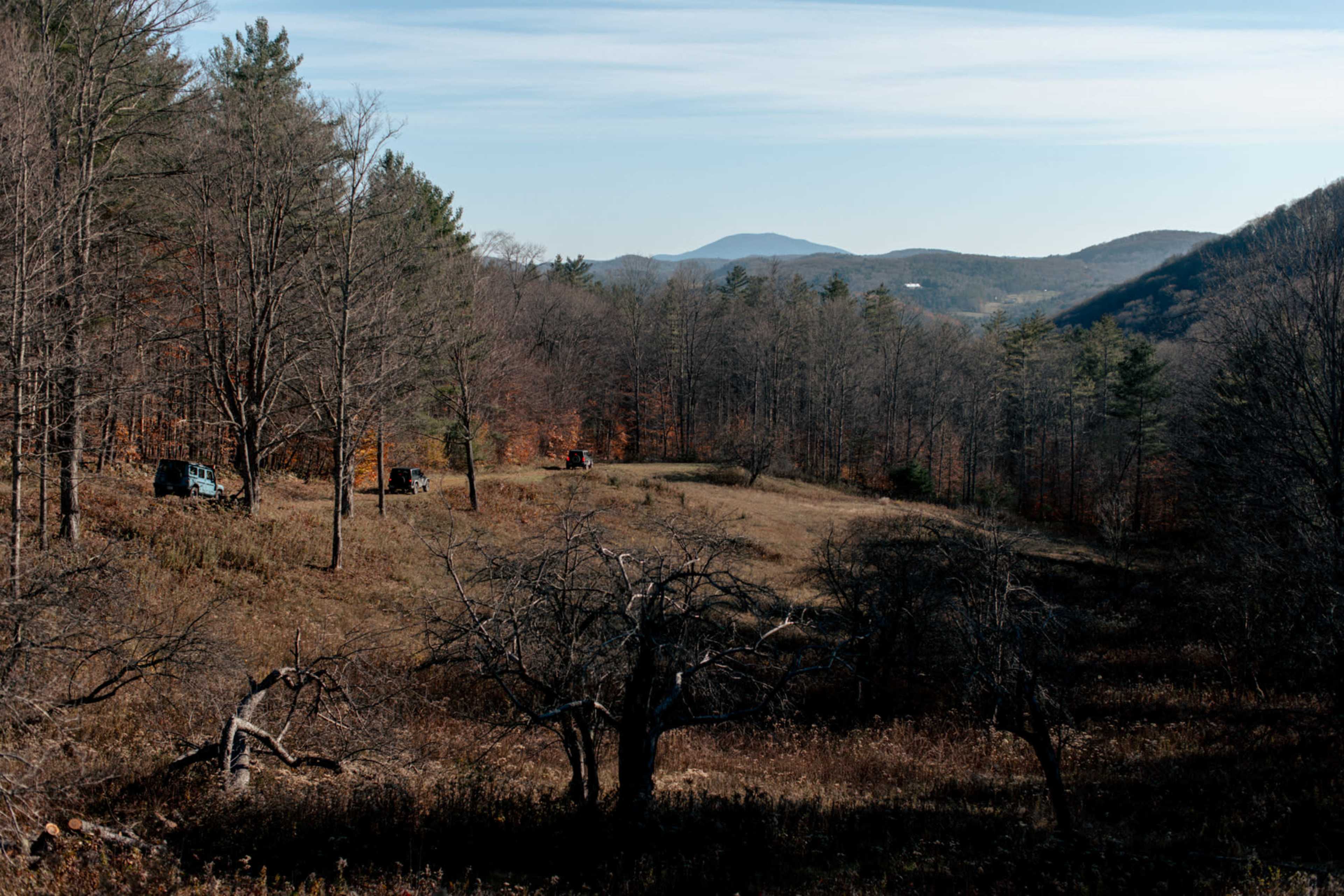 A landscape of rolling hills with sparse trees and a few vehicles parked on a grassy field under a clear blue sky.