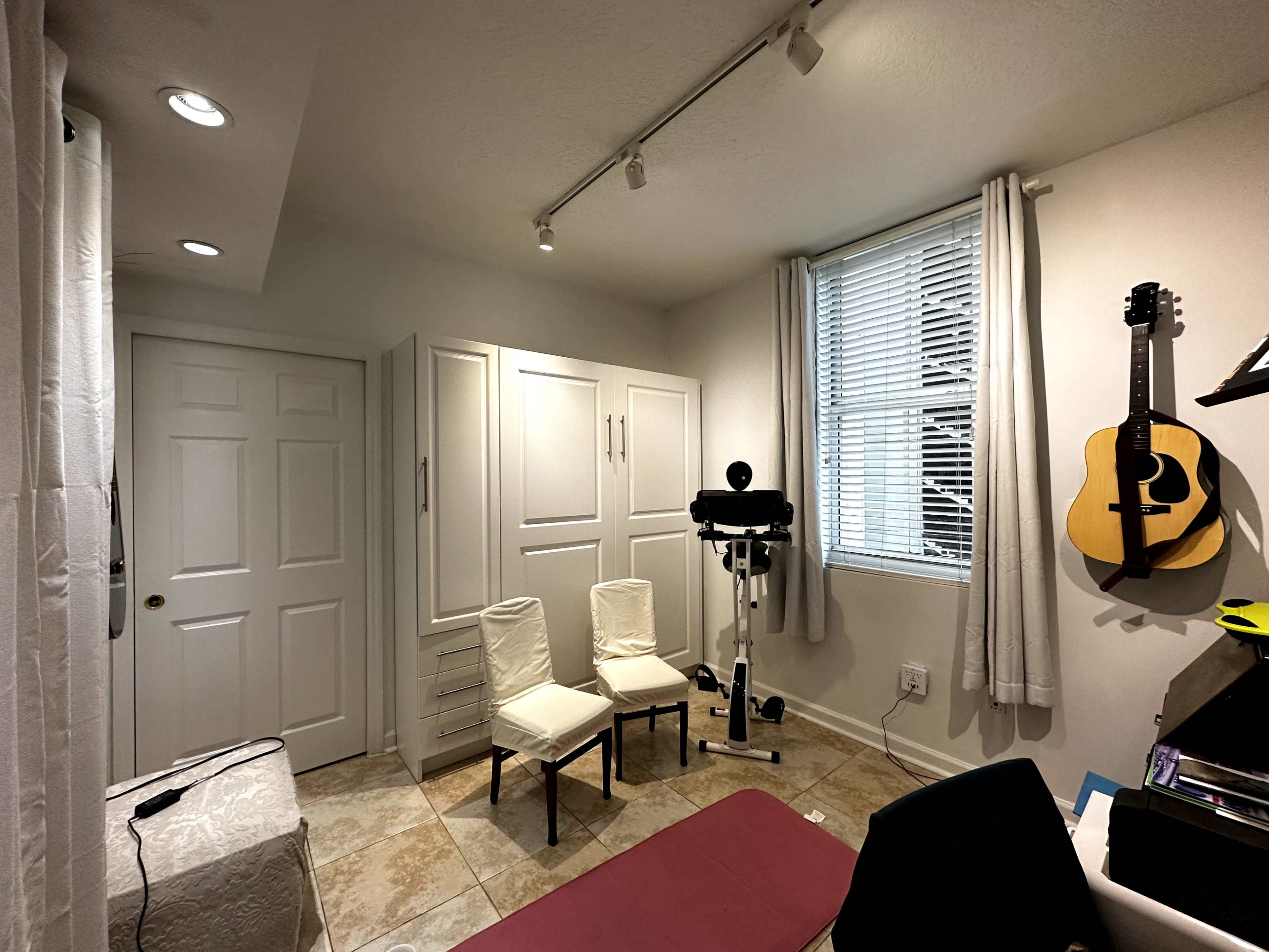 The room features two chairs, a guitar on the wall, exercise equipment, and storage cabinets.