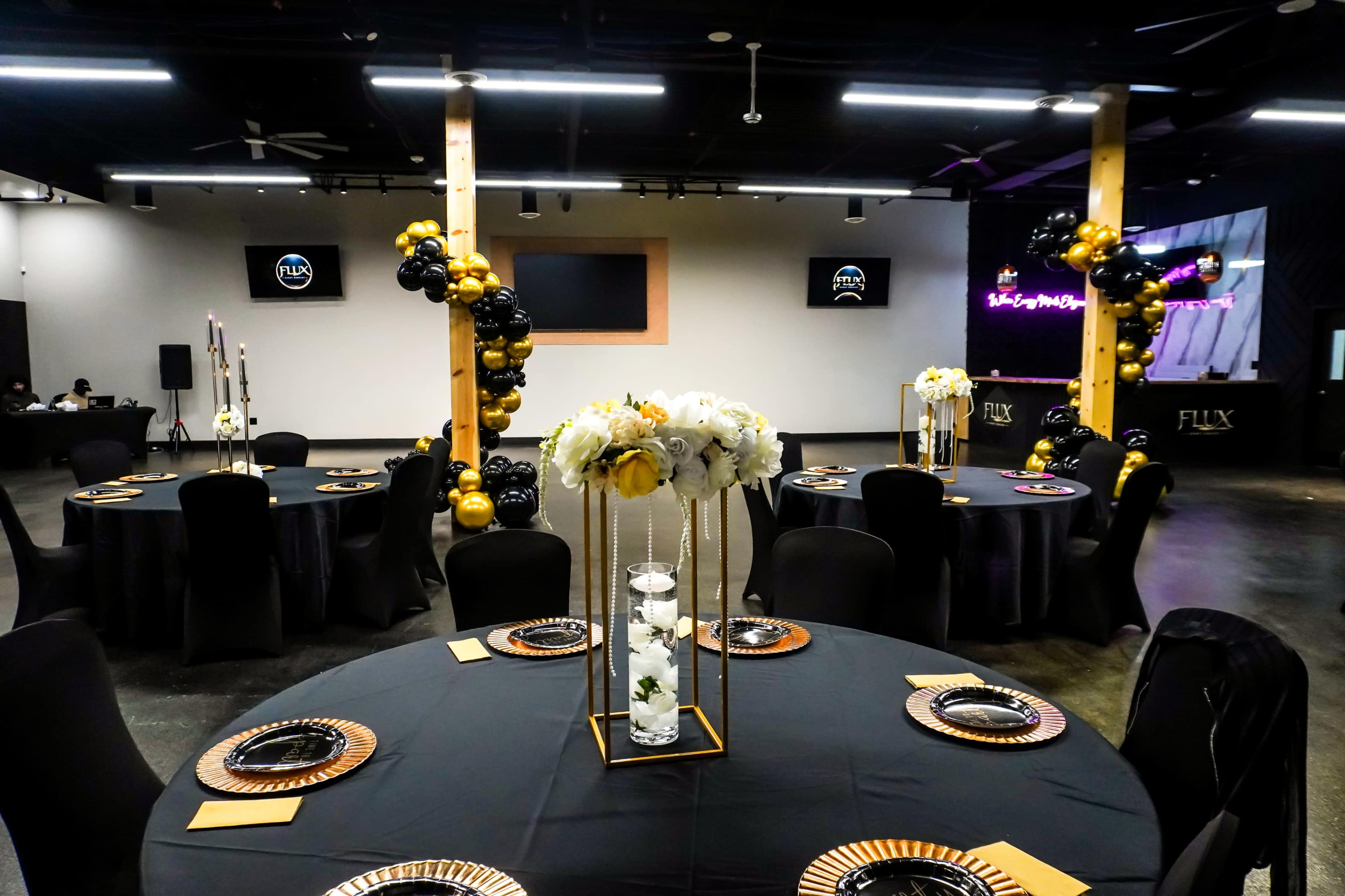 The image shows a decorated event space with round tables draped in black tablecloths, featuring floral centerpieces and gold accents, set for a formal occasion.