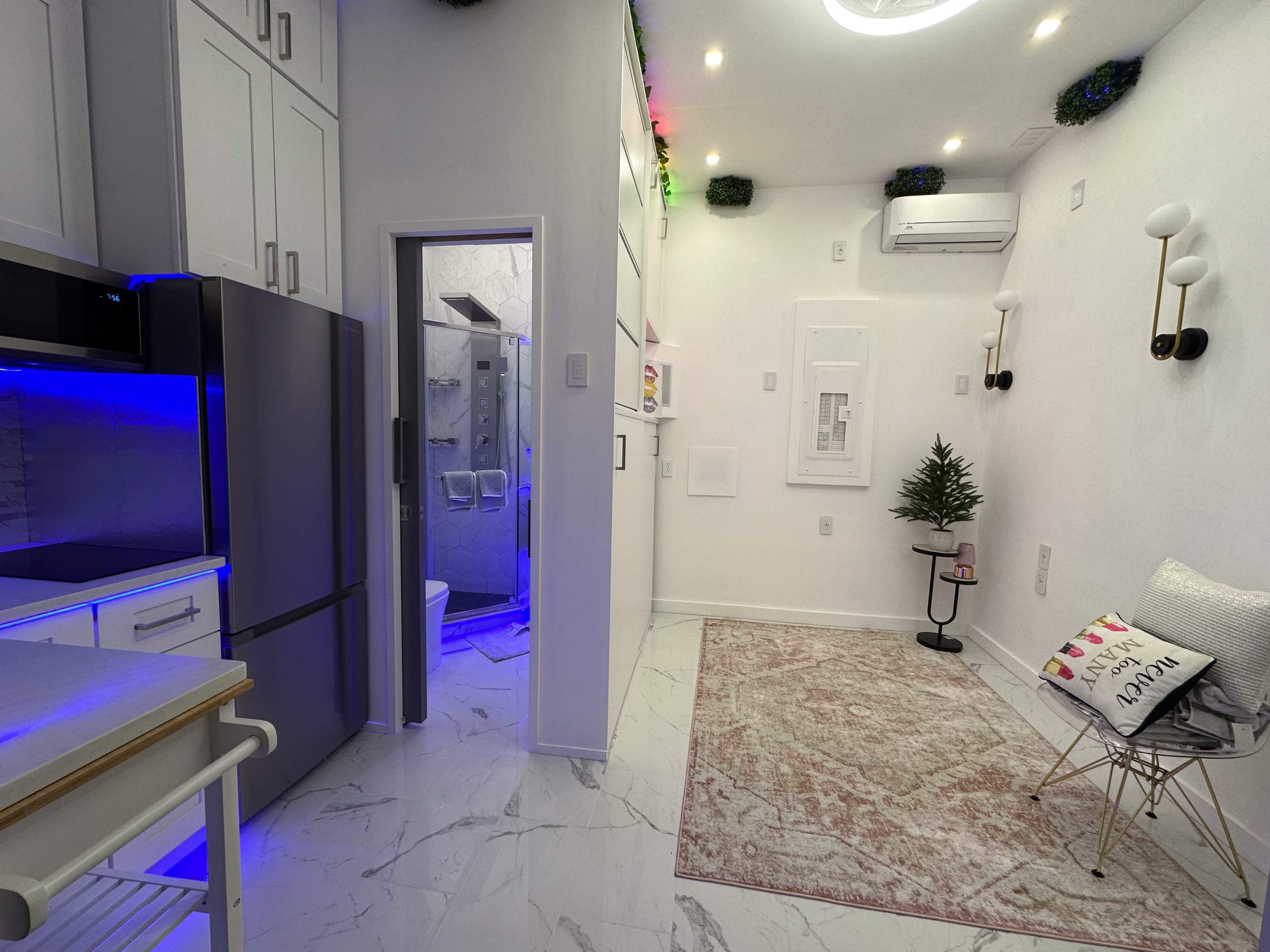 The image shows a modern, compact kitchen and living area featuring sleek cabinetry, a refrigerator, a small dining table, and a bathroom visible through an open door, all with contemporary lighting and decor.