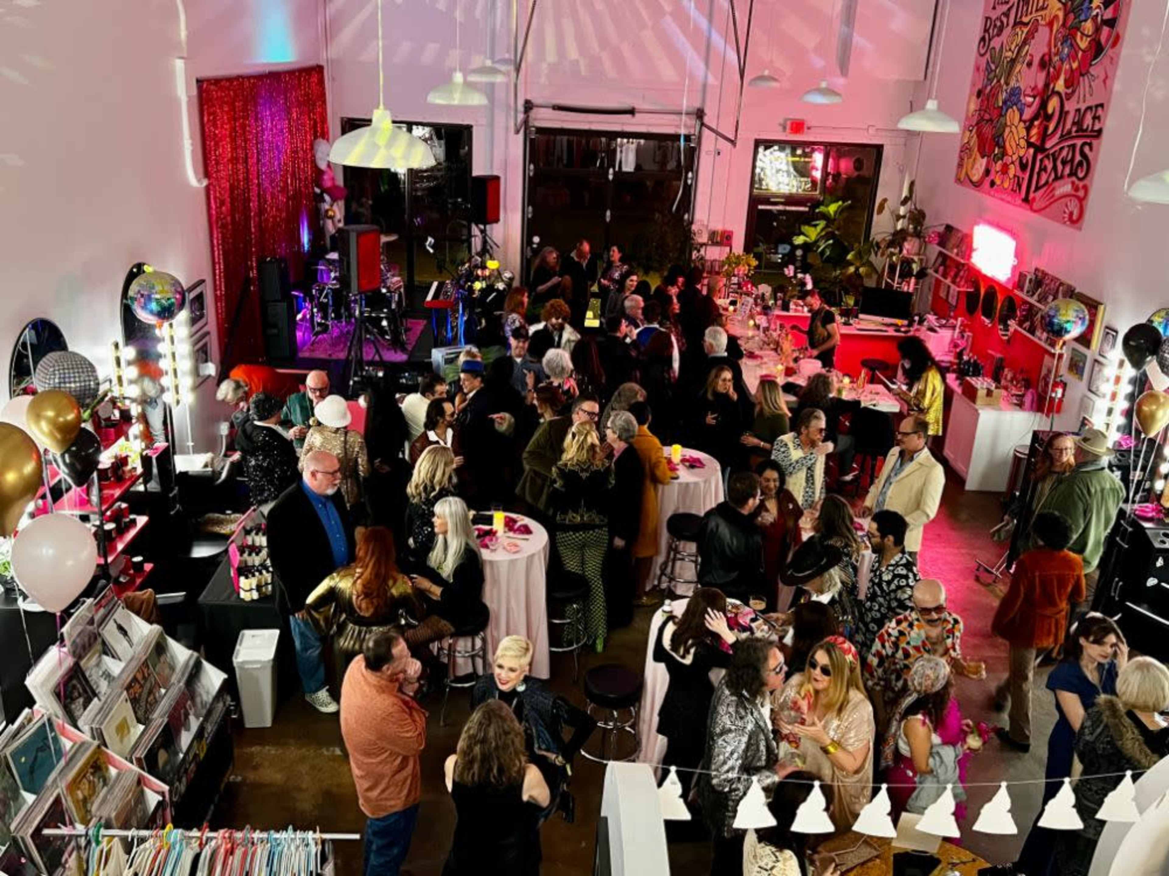 The image shows a lively party scene inside a spacious venue, filled with people socializing, colorful decorations, and a stage setup for live music.