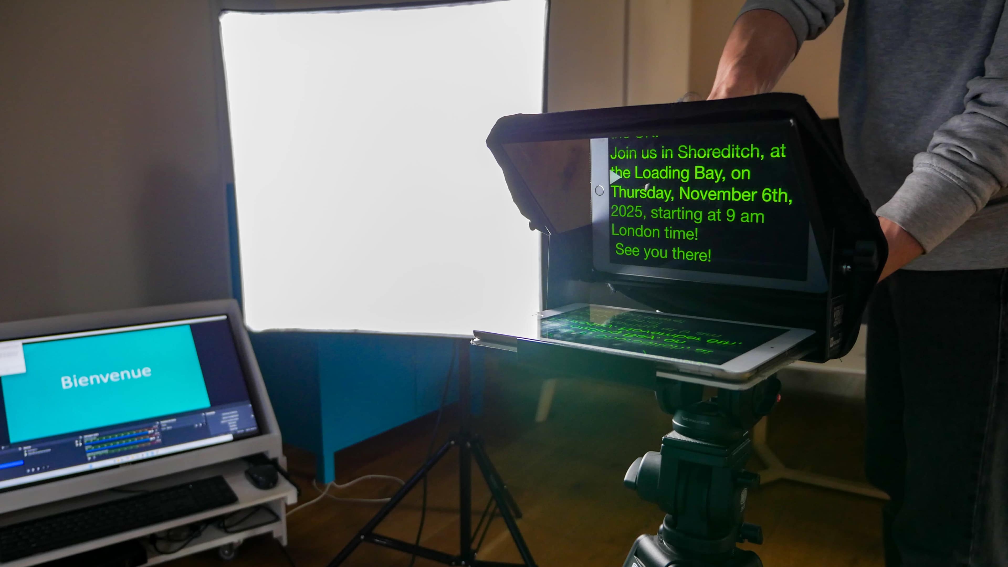A person operates a teleprompter next to a lit backdrop and a computer screen displaying "Bienvenue."