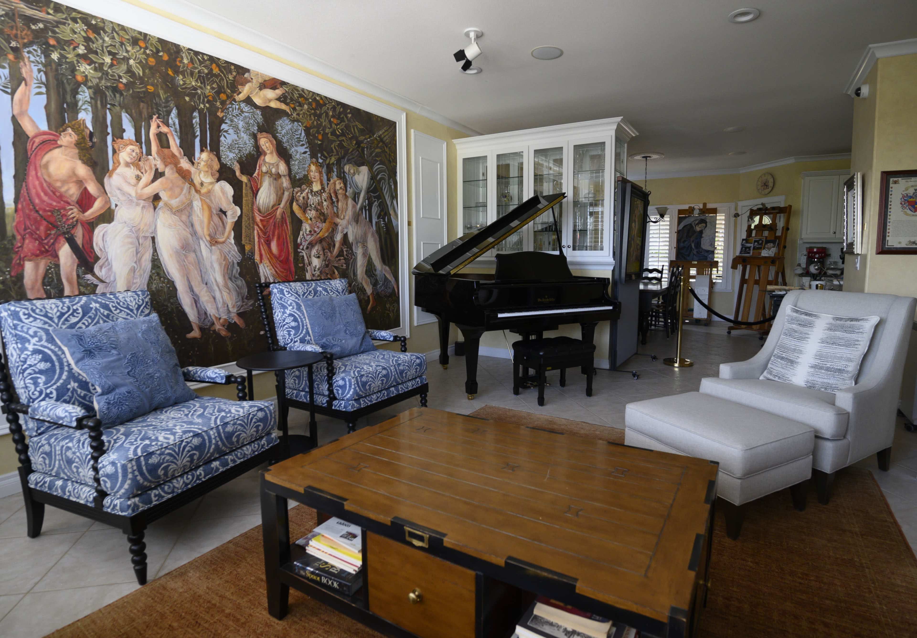 A living room with blue patterned chairs, a white armchair, a wooden coffee table stacked with books, a black grand piano, and a large mural of classical figures on the wall.