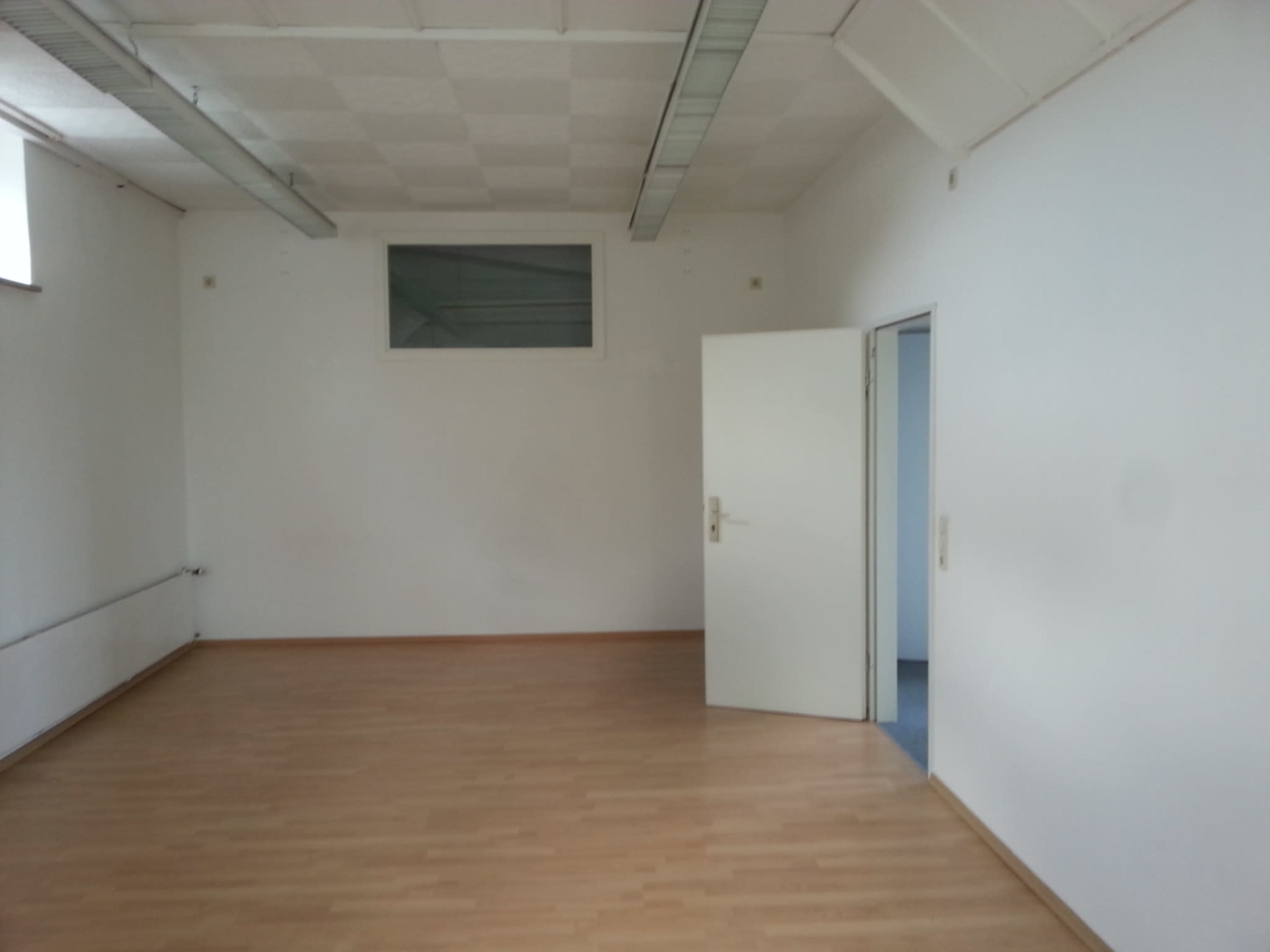 The image shows an empty room with wooden flooring, white walls, a window, and an open door.