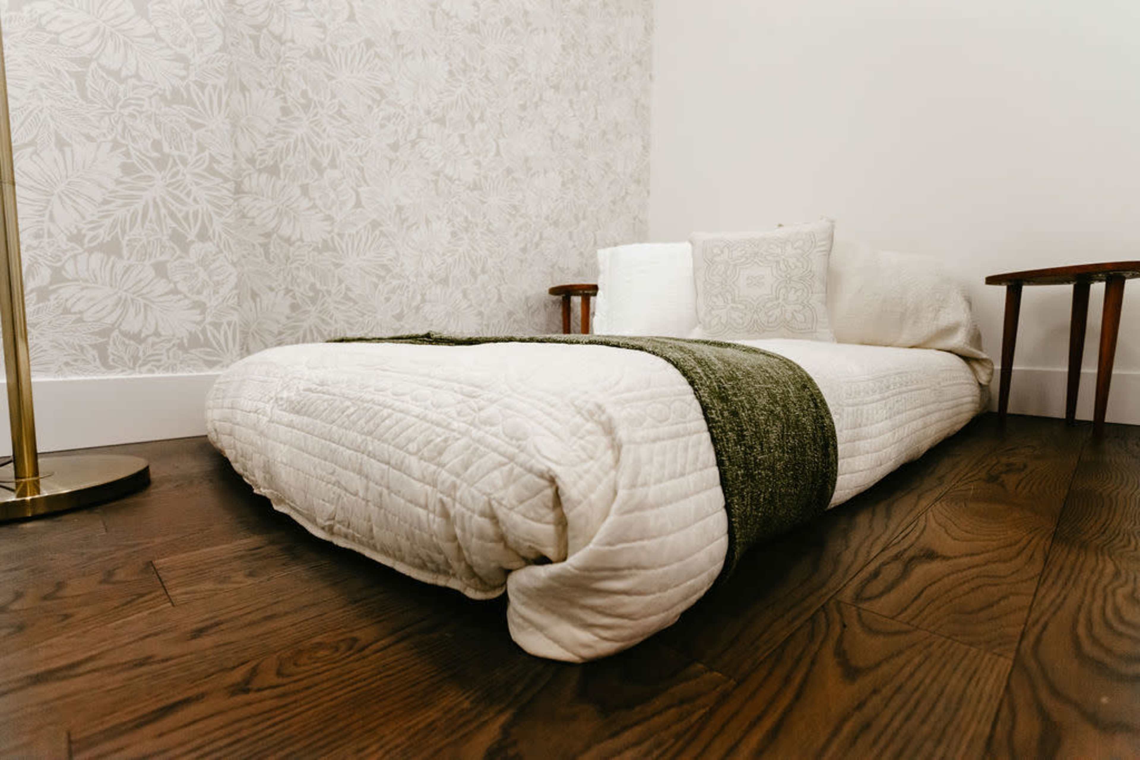 A low bed with a white quilt and a green throw is positioned against a patterned wall, accompanied by two wooden side tables.