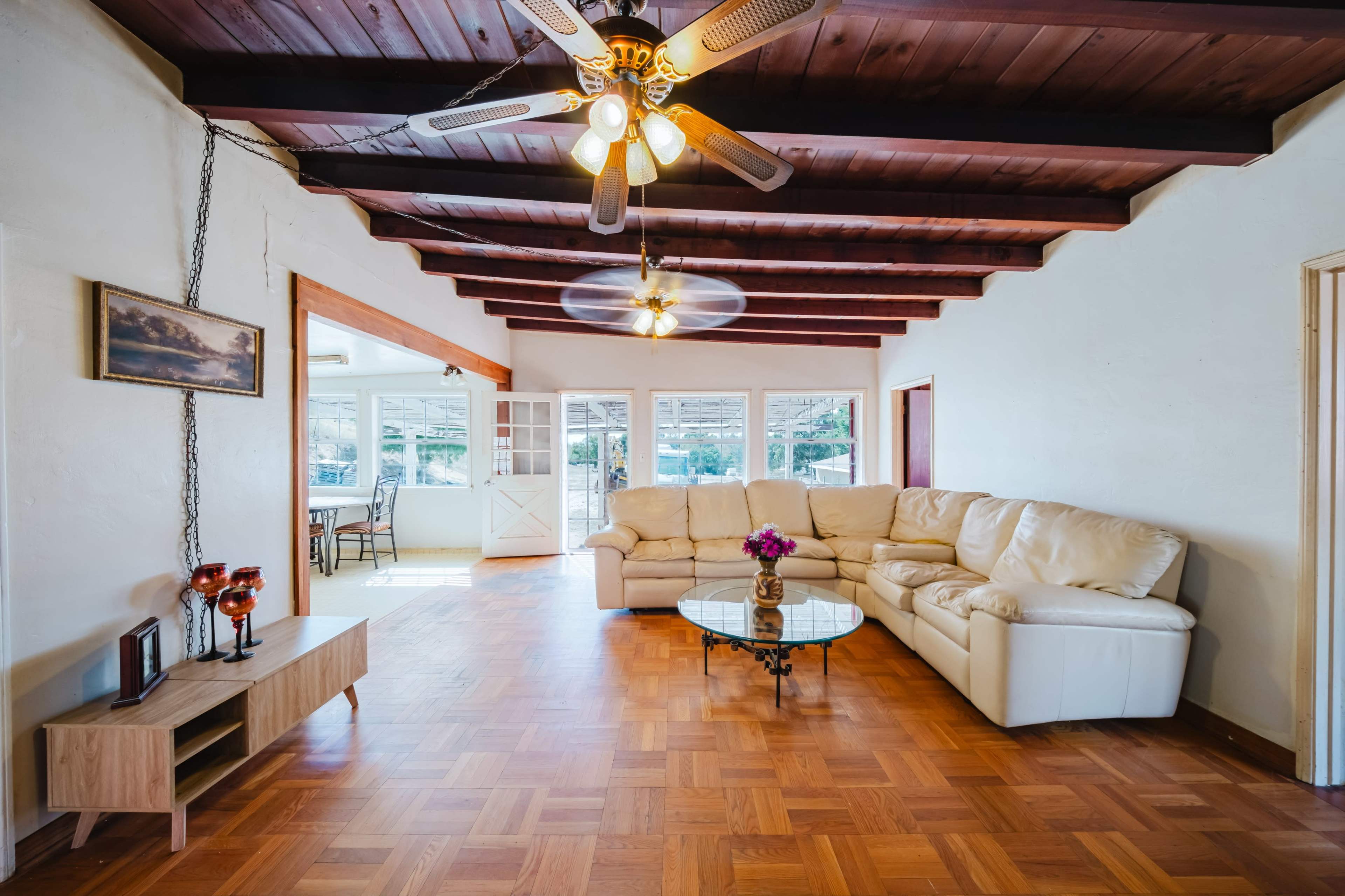 The image shows a spacious living room with a large sectional couch, a glass coffee table, and wooden ceiling beams, leading to a sunlit area with glass doors.