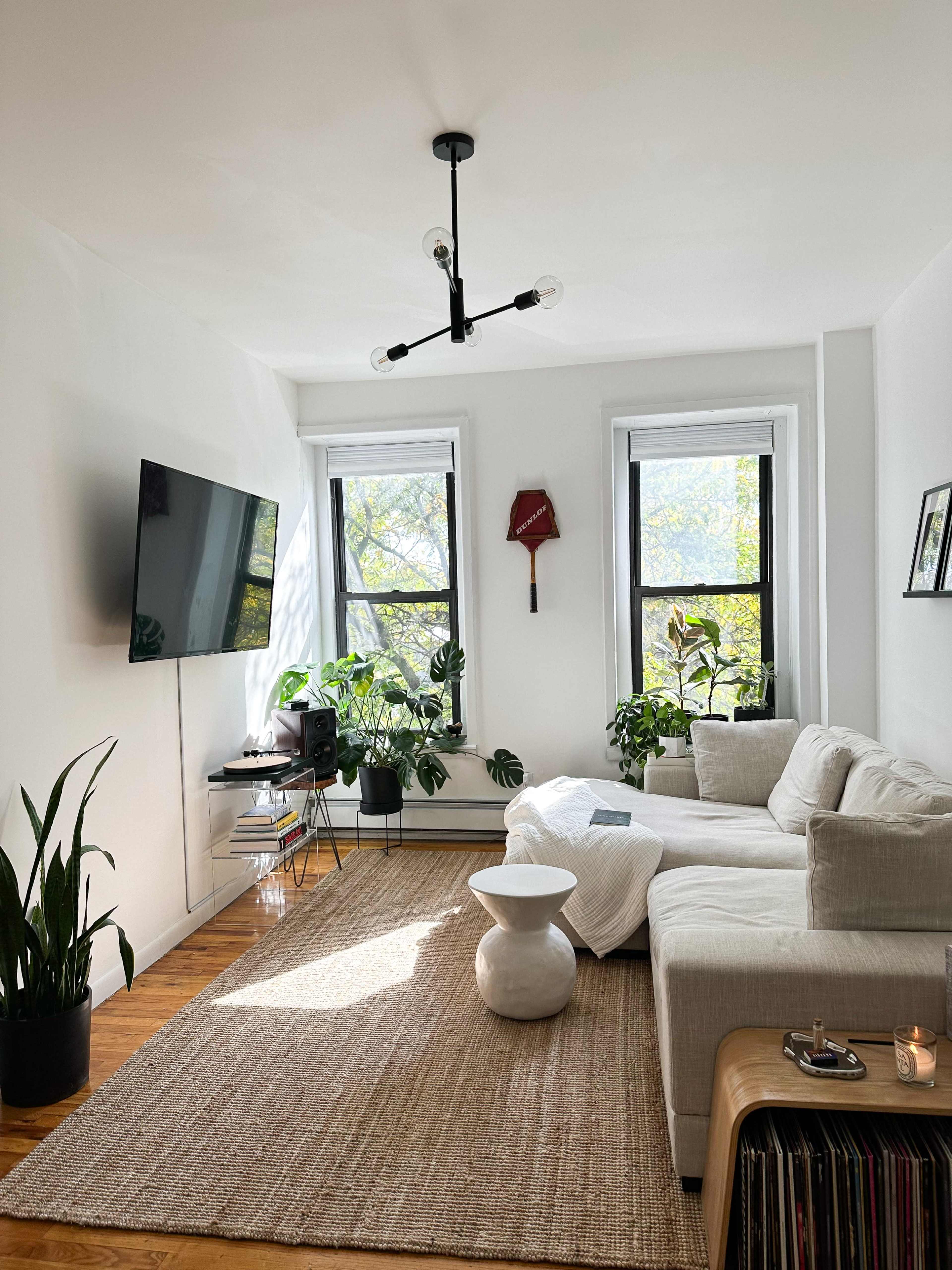 A bright living room features a sectional sofa, a television on the wall, large windows with plants, and a round coffee table.