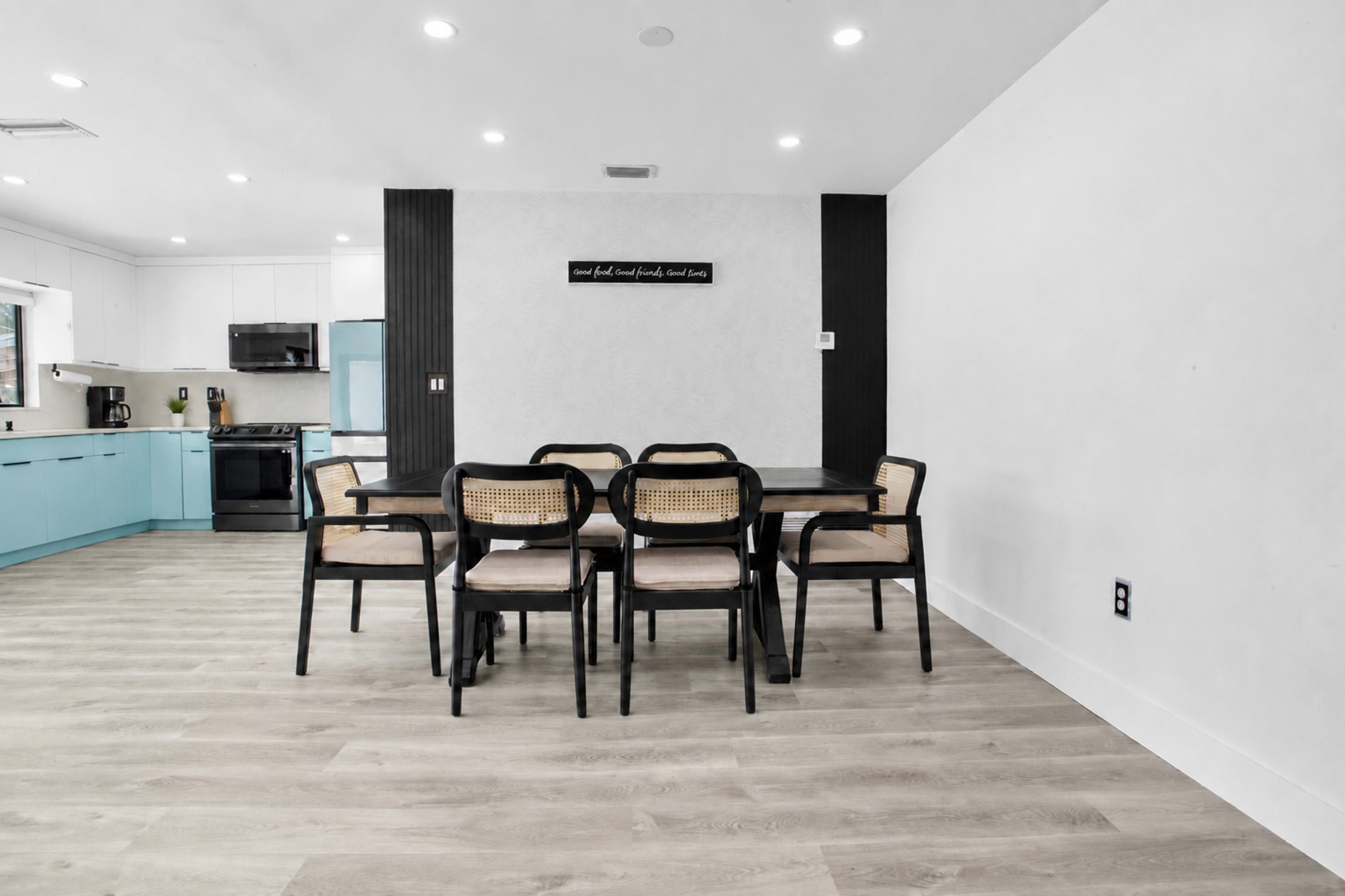 A modern dining area features a large black table surrounded by six chairs, with a kitchen in the background displaying light blue cabinets and white appliances.