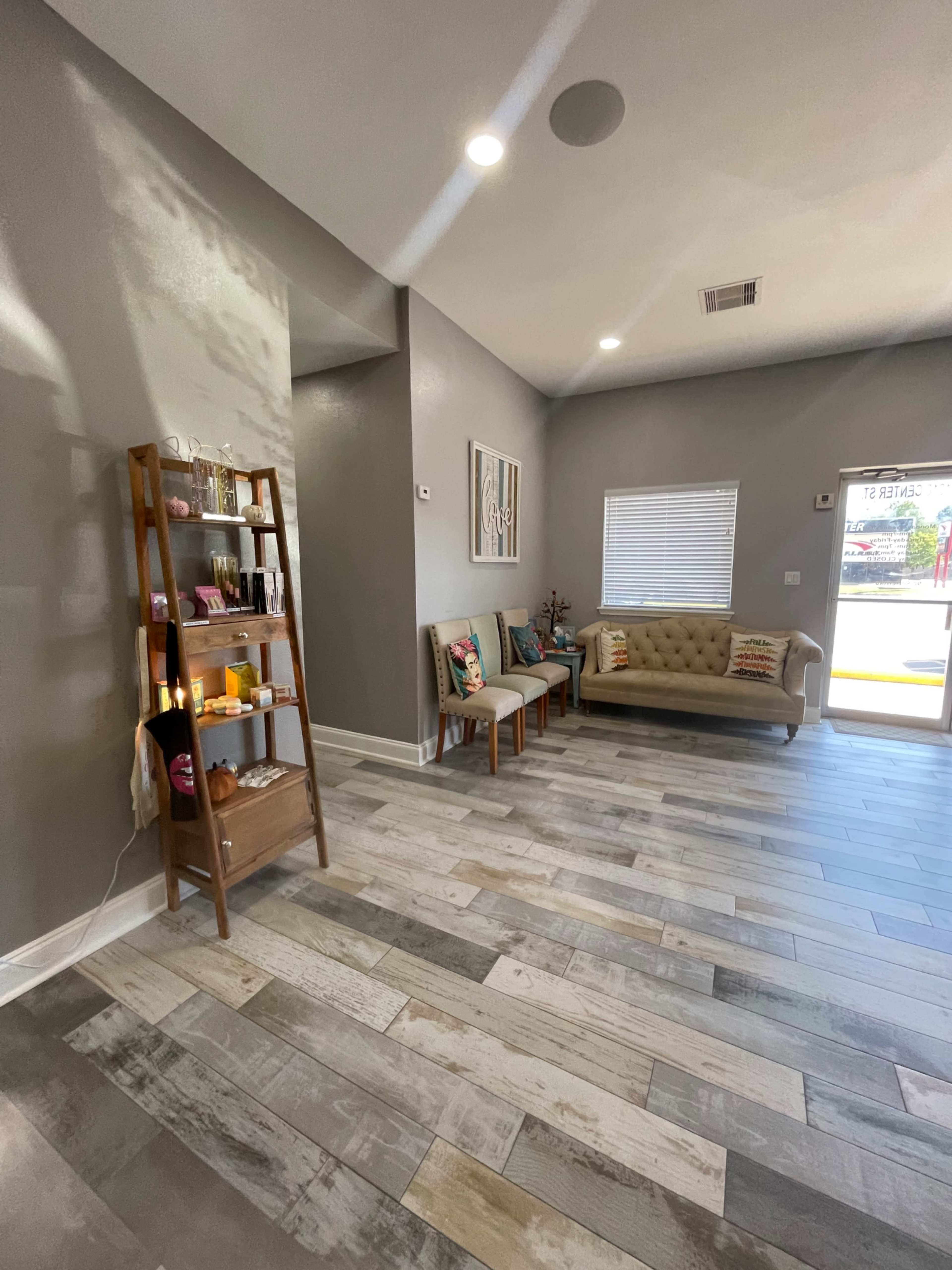 A modern interior space with a light gray wall, wooden flooring, a beige sofa, two wooden chairs, and a shelving unit displaying various products.