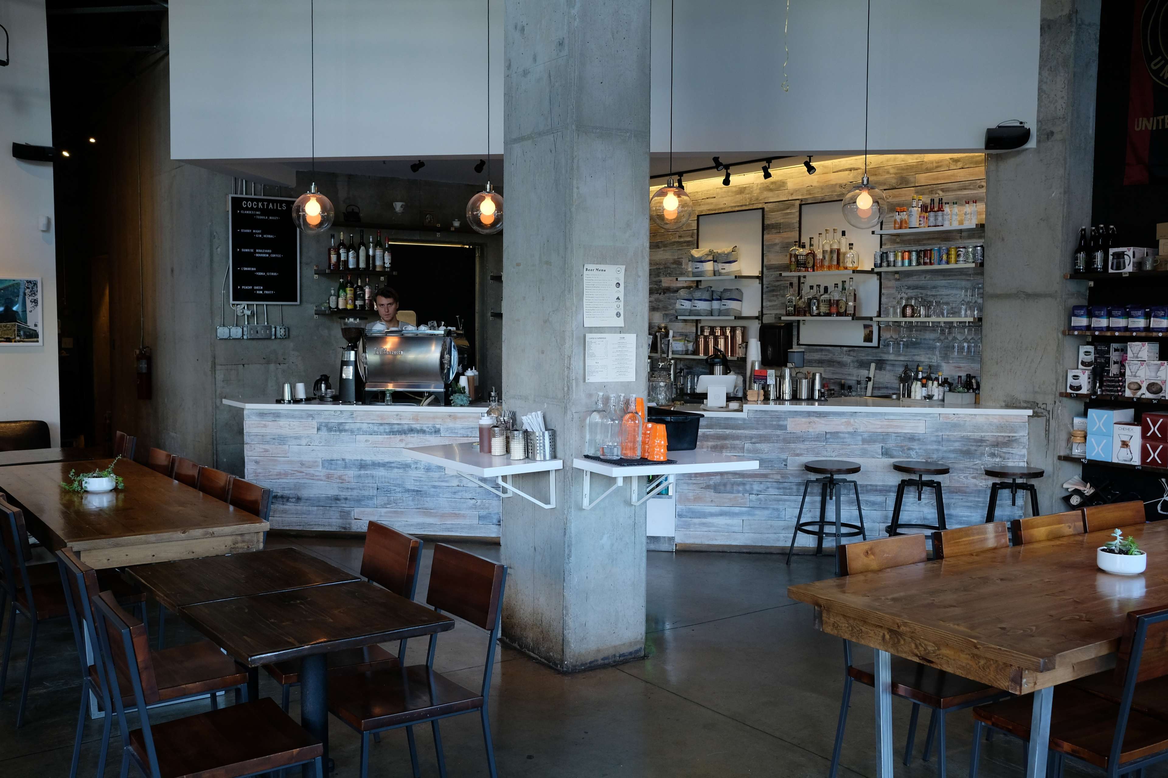 A modern café interior with a service counter, shelves of beverages, and wooden tables arranged around the space.