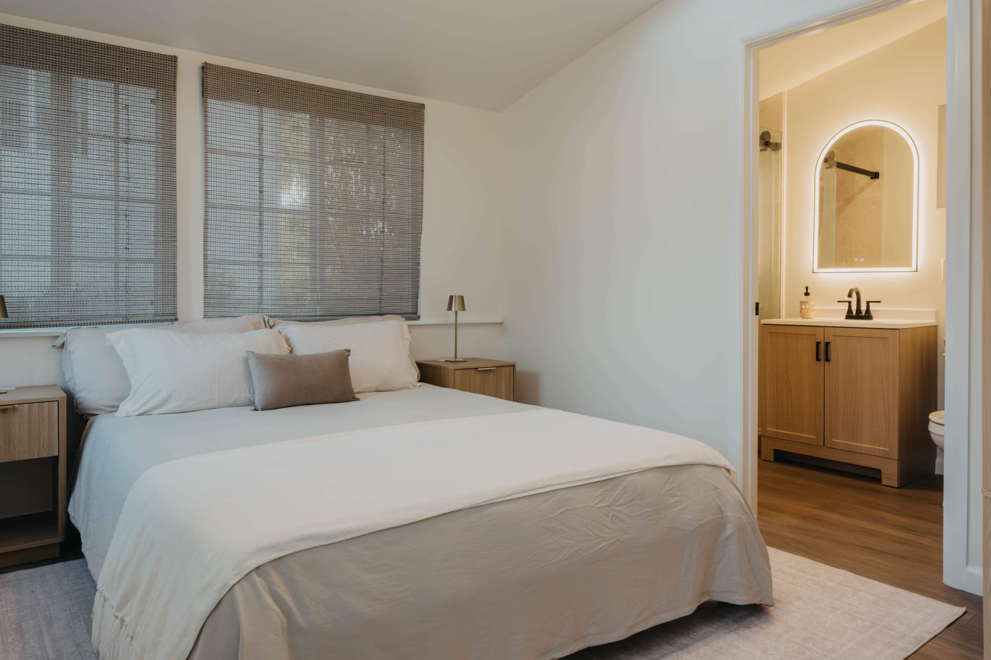 The image shows a minimalist bedroom featuring a bed with a light-colored bedspread, two bedside tables, and an open bathroom with a mirror and sink visible in the doorway.