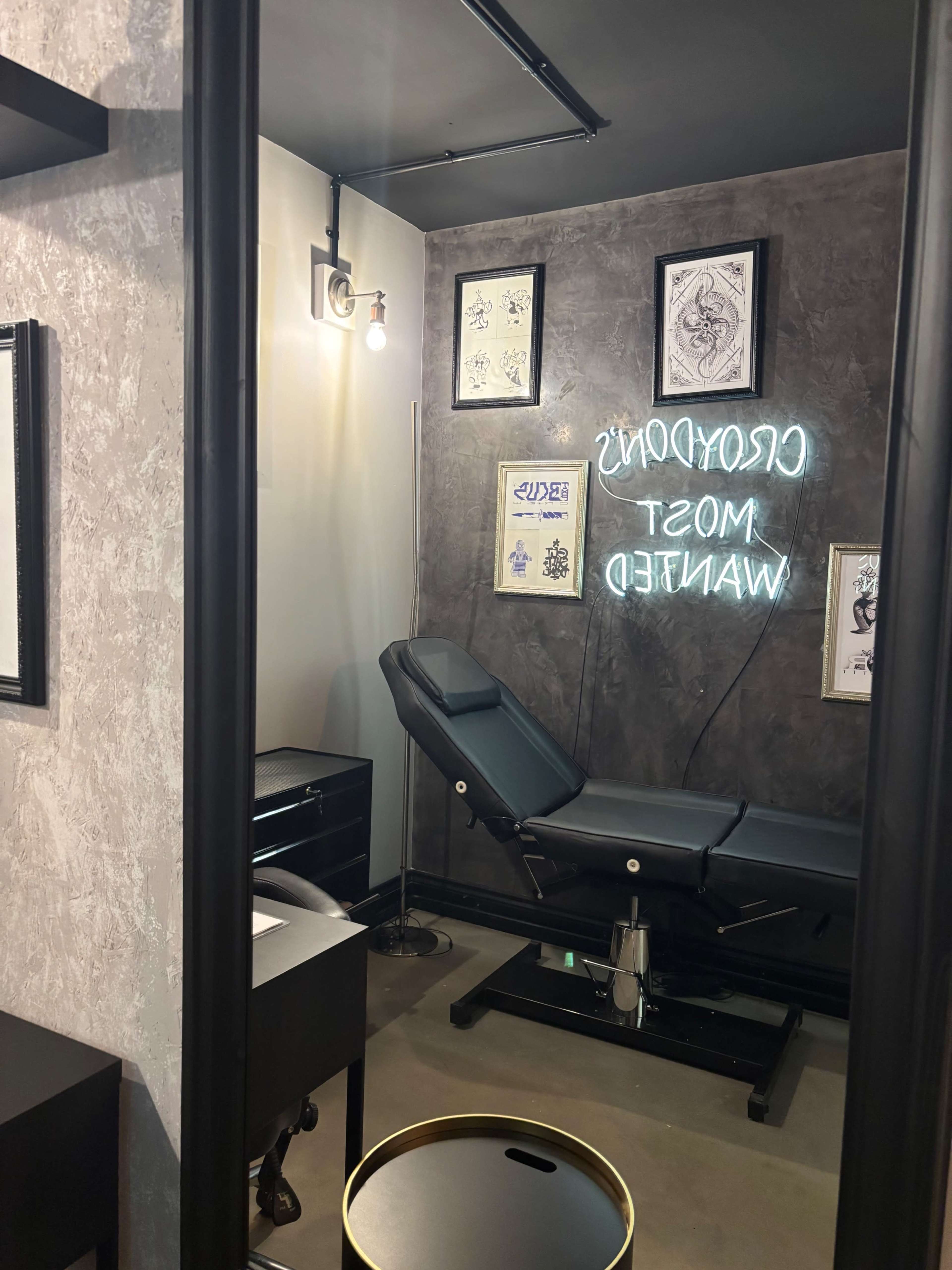 The image shows a tattoo studio featuring a tattoo bed, neon signage with the text "CROOKED TATTOOS MOST WANTED," and framed artwork on the walls.