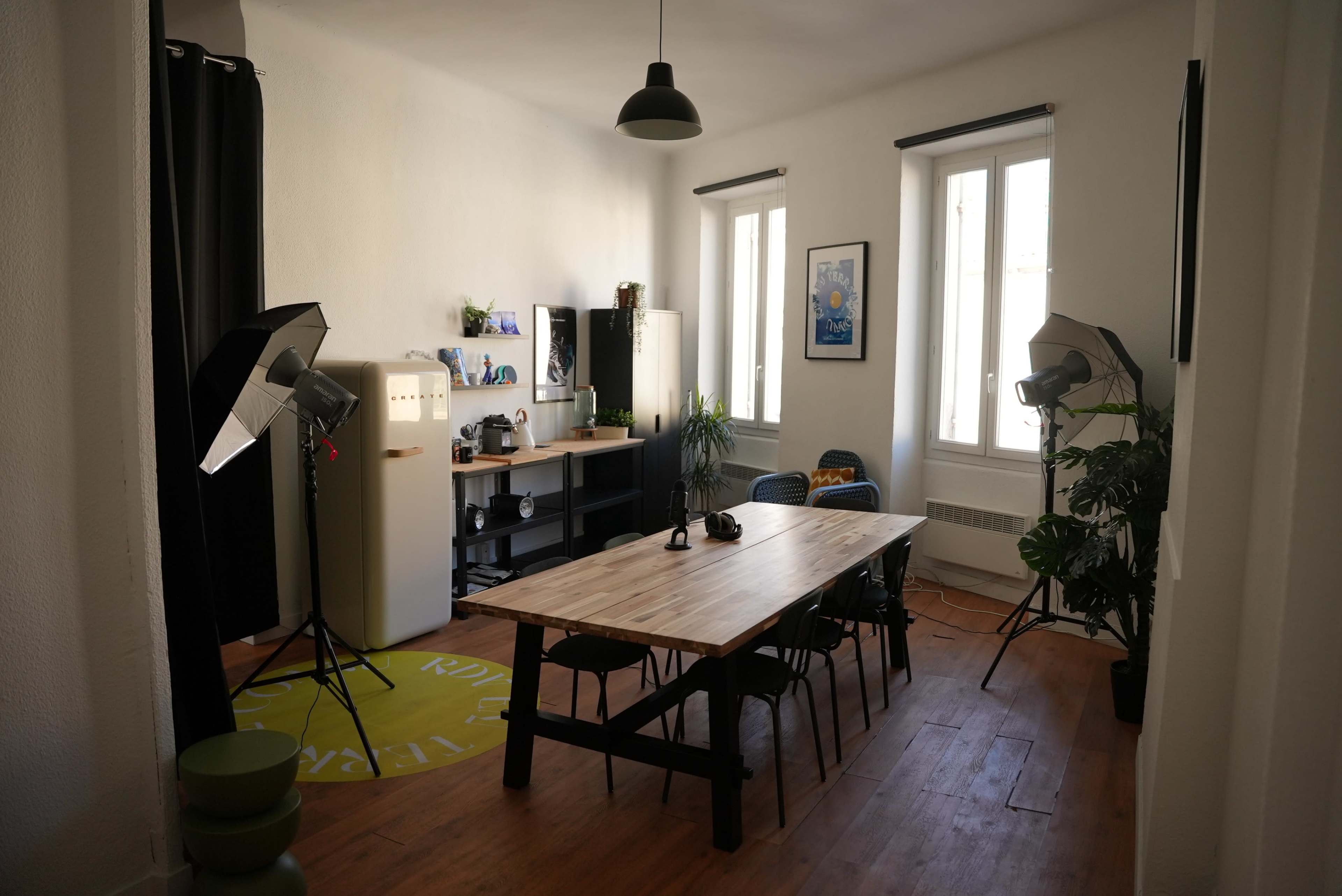 A well-lit room features a wooden table surrounded by chairs, with photography lighting equipment set up in the corners and decorative plants along the walls.