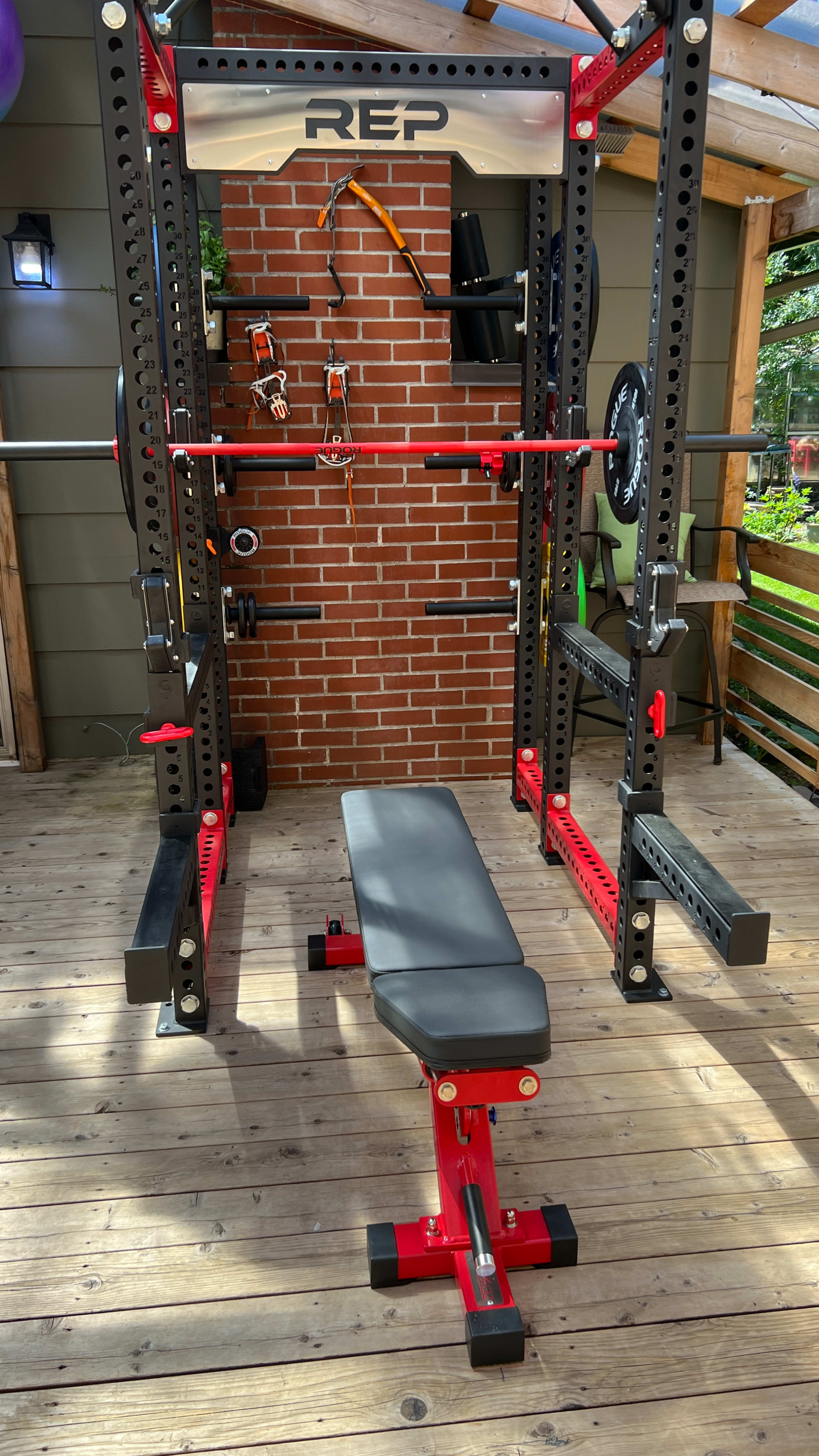 A weightlifting setup with a squat rack, bench, and various accessories against a brick wall in a wooden structure.