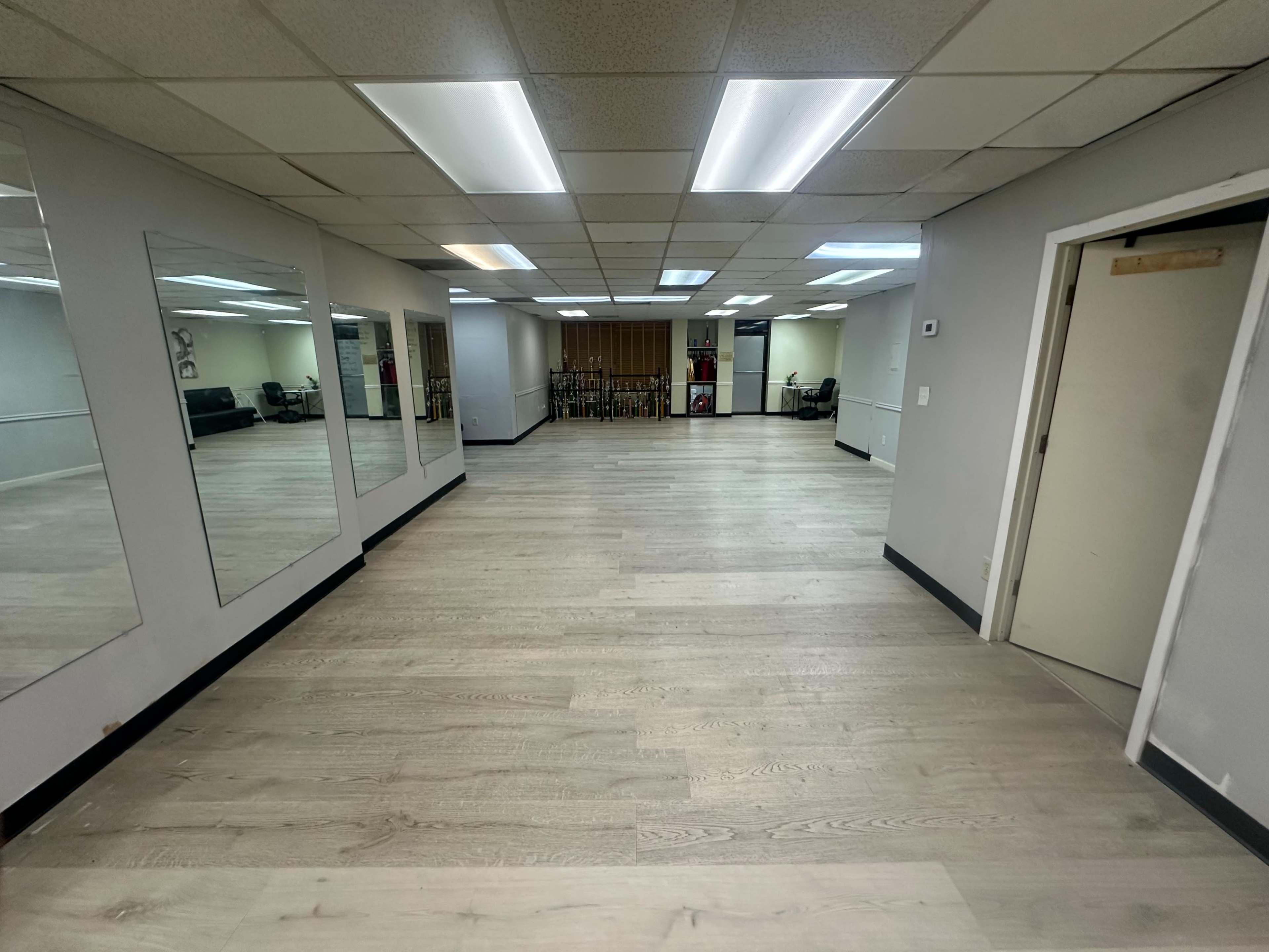 The image shows an empty room with wooden flooring, bright overhead lights, and mirrors on one wall.