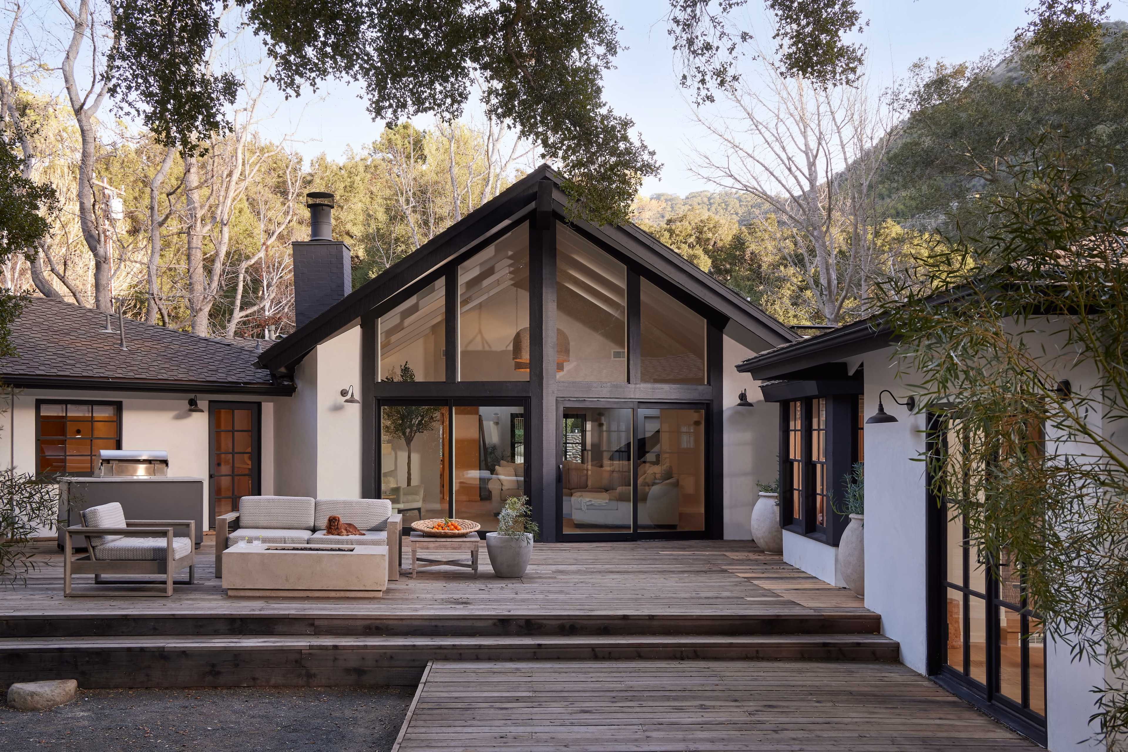 The image shows a modern home with a wooden deck, large glass windows, and a gabled roof, set amidst trees and natural surroundings.