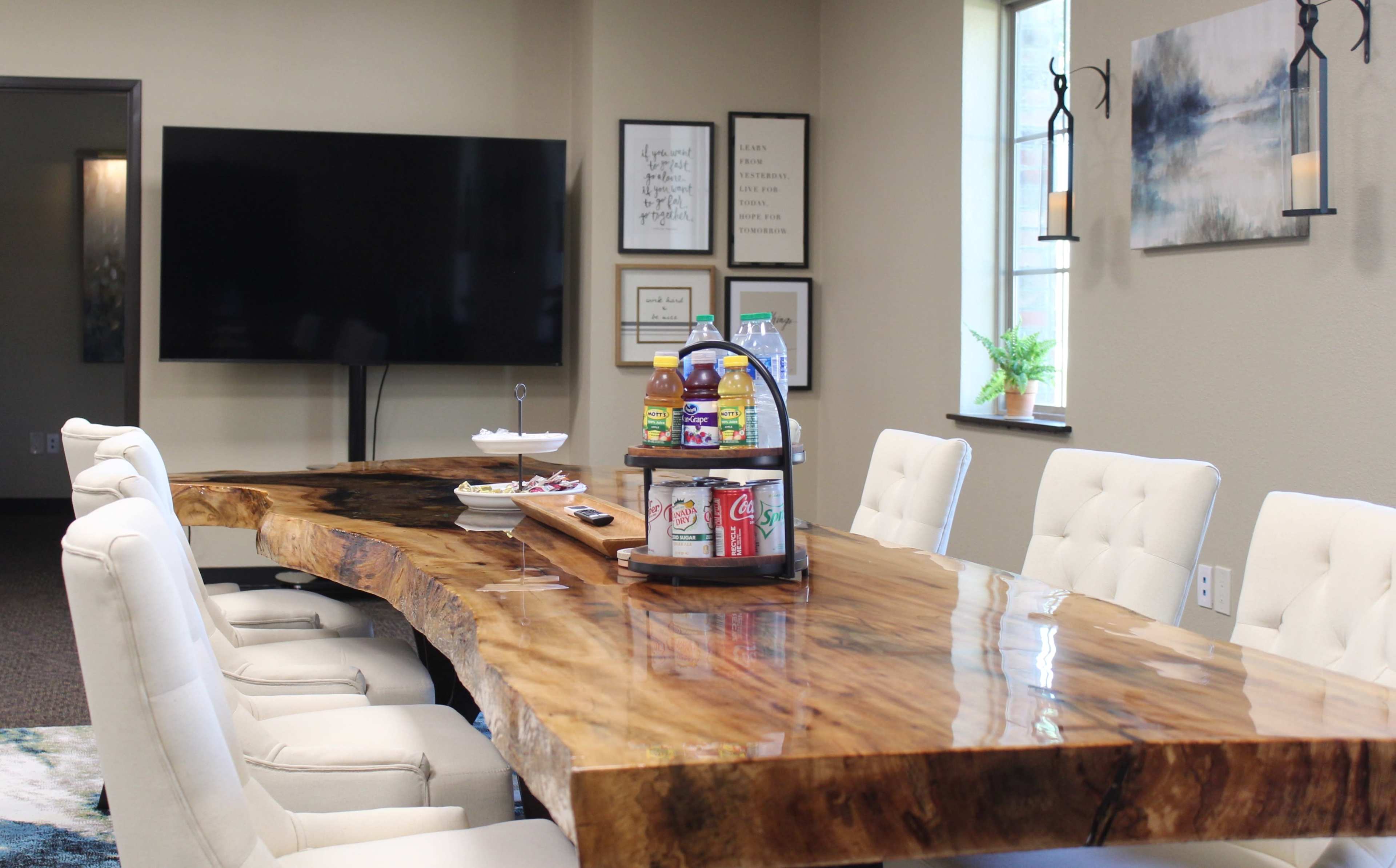 A large, polished wooden table with a circular serving tray that holds drinks, surrounded by white upholstered chairs in a well-lit room.