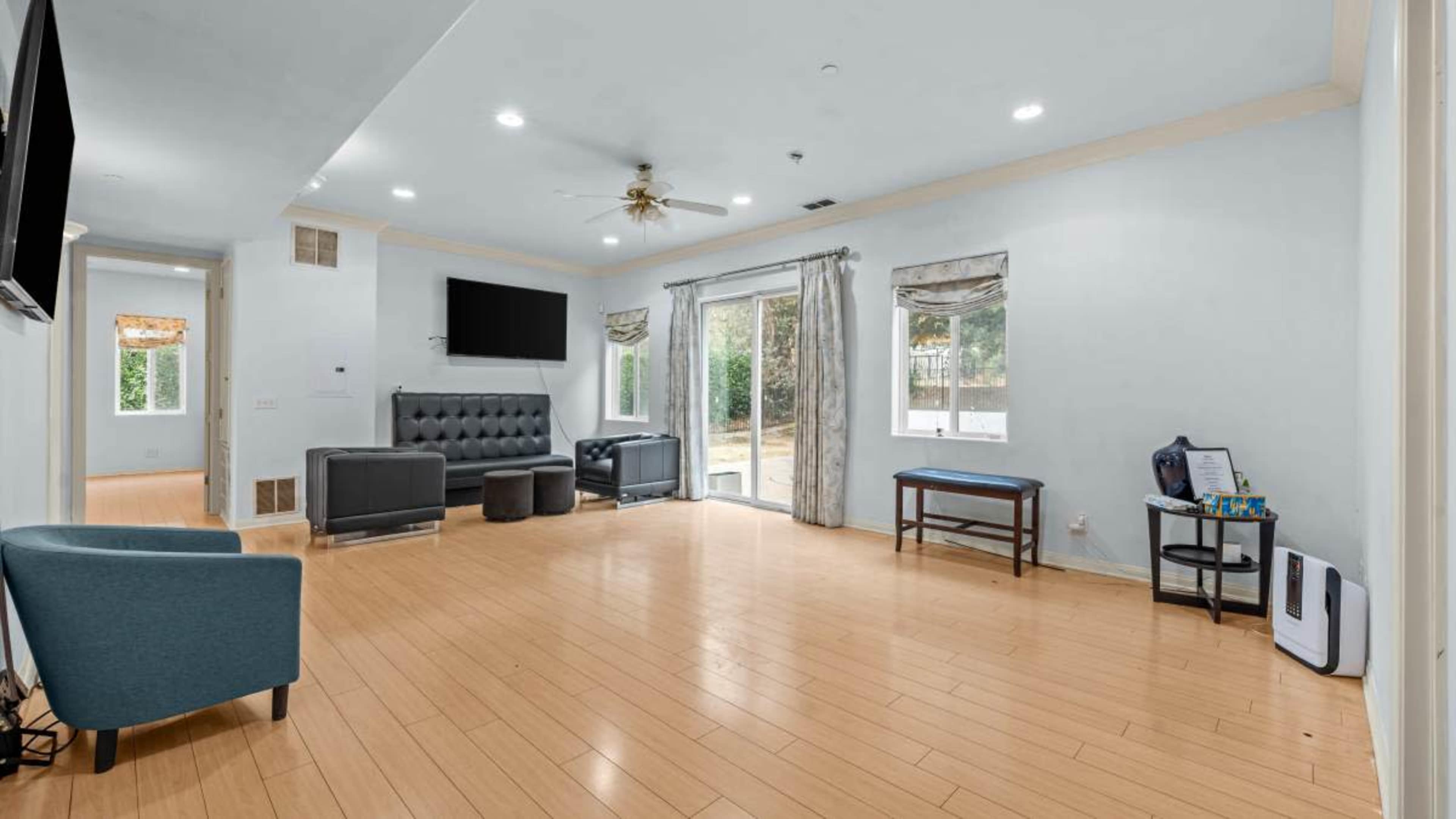 The room features light blue walls, wooden flooring, a seating area with a sectional sofa and chairs, a sliding door leading to an outdoor space, and wall-mounted televisions.