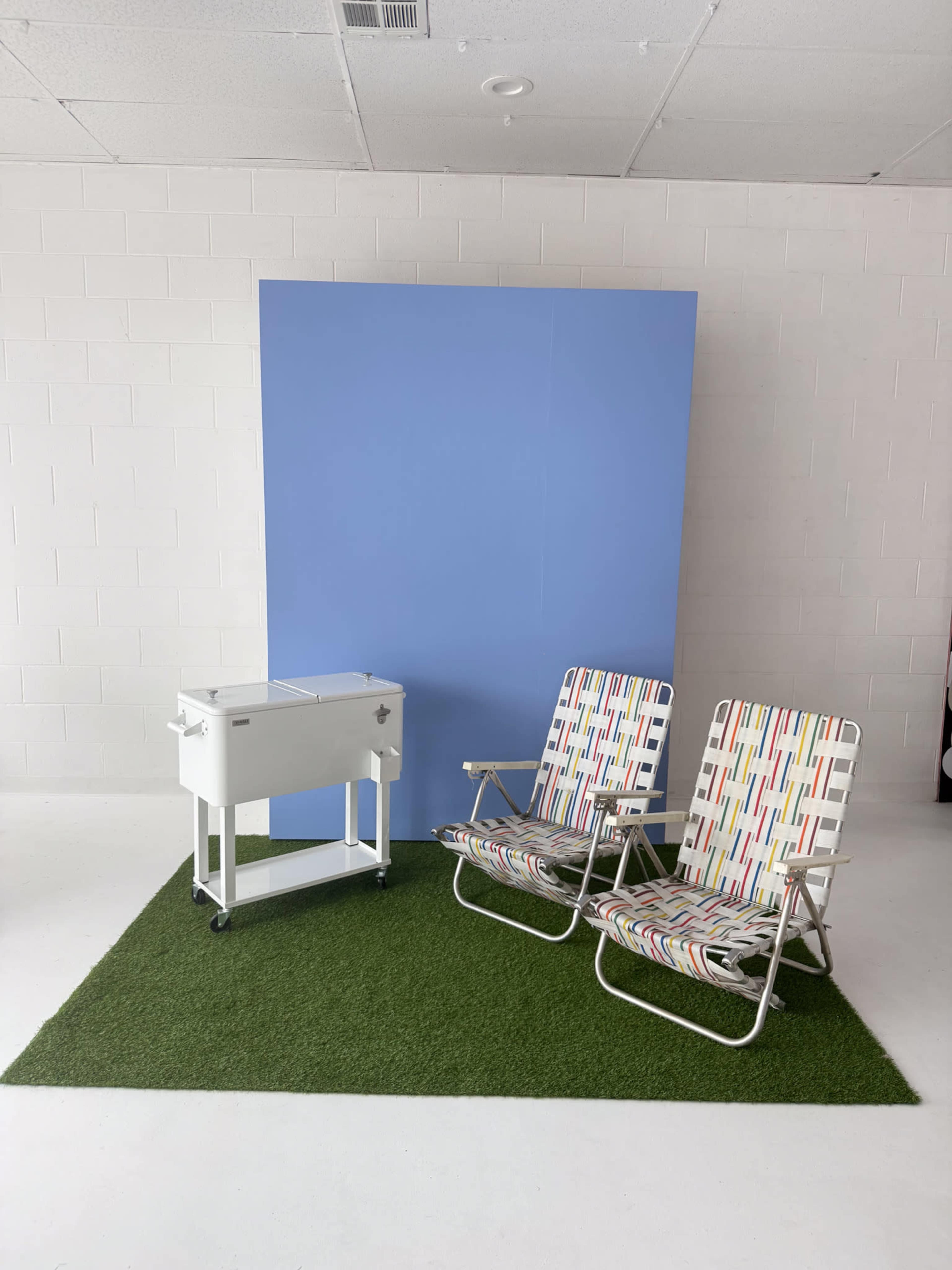 The image shows two folding lounge chairs with a colorful pattern, positioned next to a portable cooler on wheels, against a blue backdrop with a patch of artificial grass on the floor.