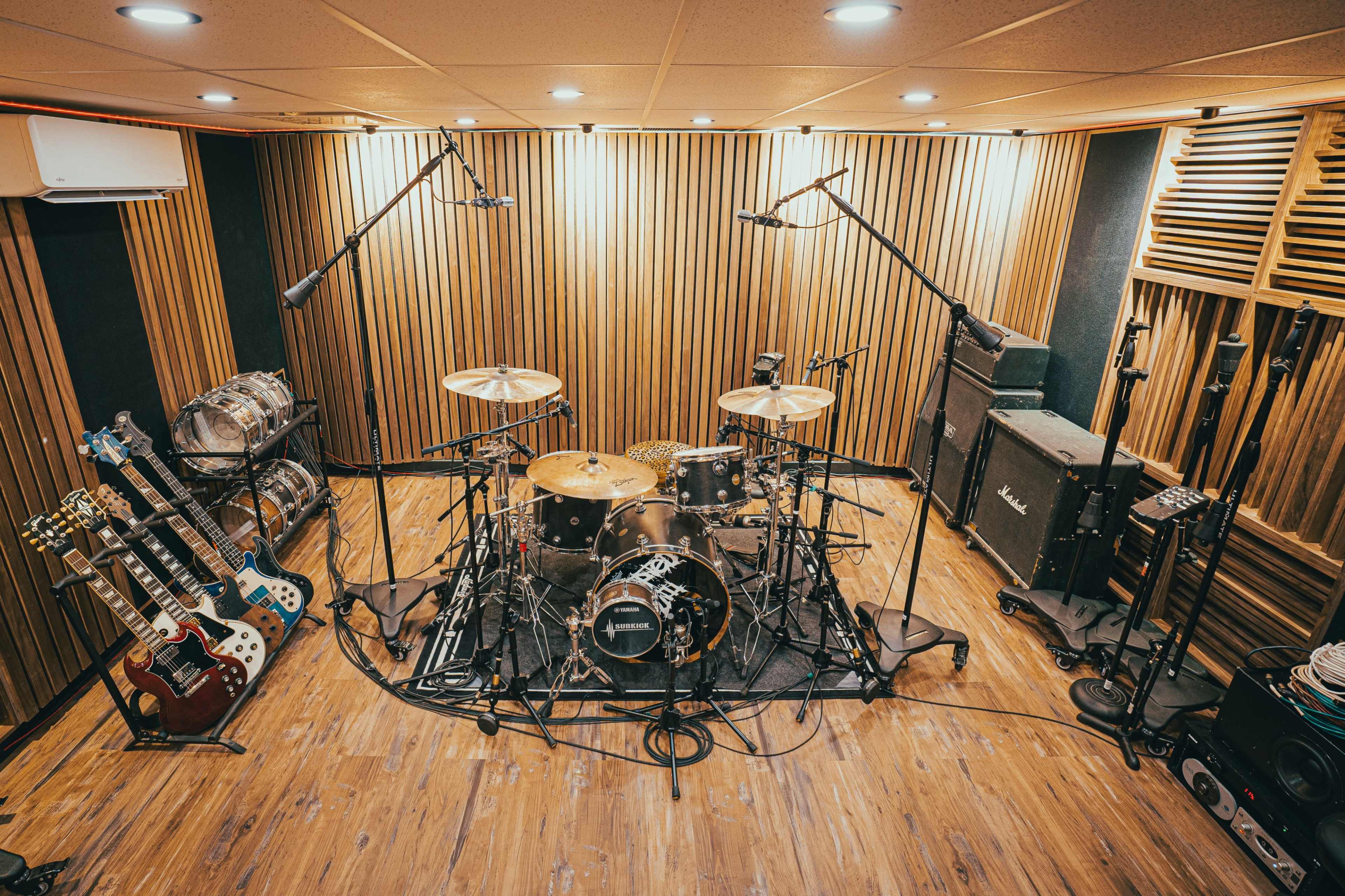 The image shows a music studio with a drum set positioned centrally, surrounded by guitars, amplifiers, and microphones.