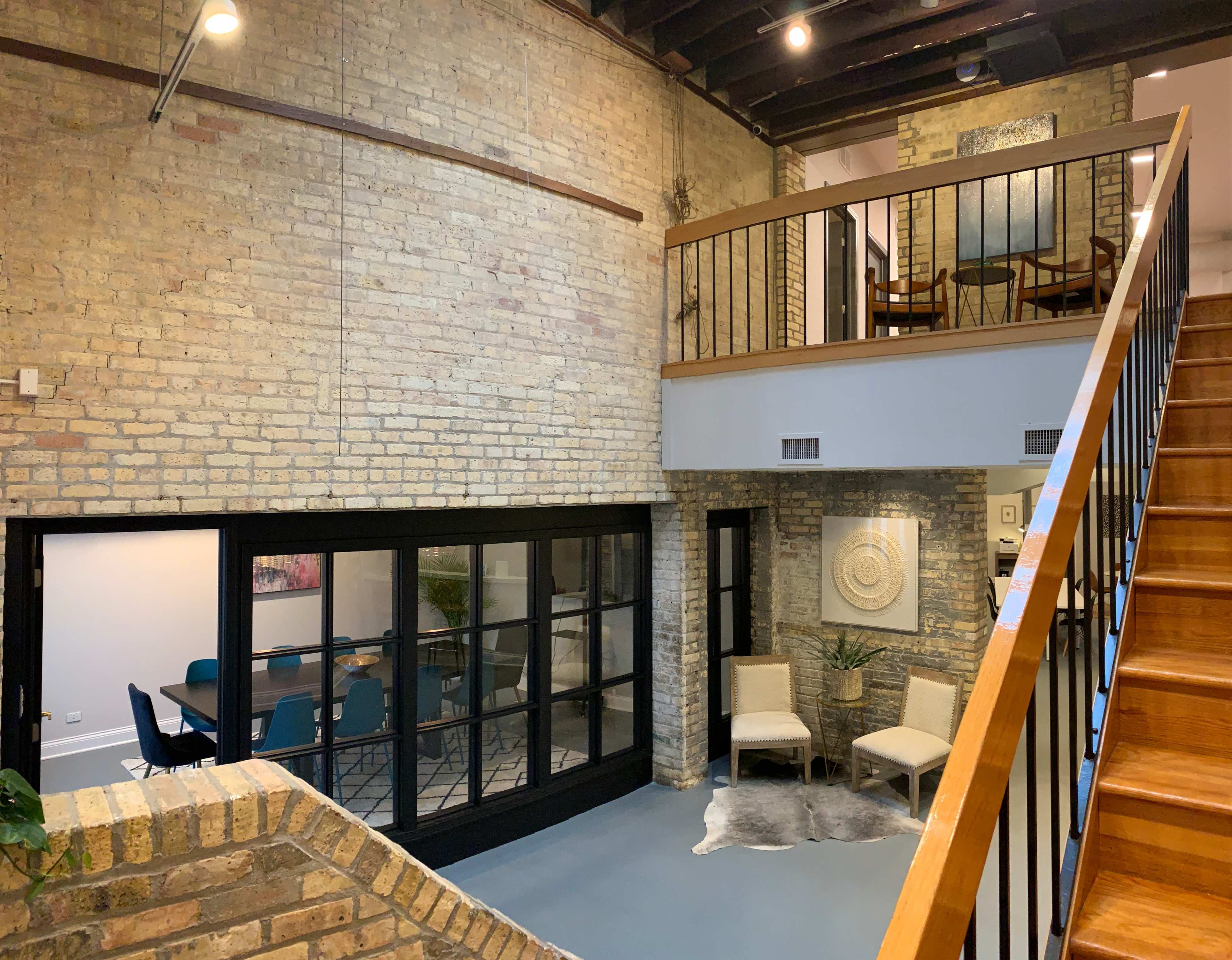 The image showcases a modern interior space with exposed brick walls, a wooden staircase leading to a balcony, and a glass door opening to a room with a dining table.