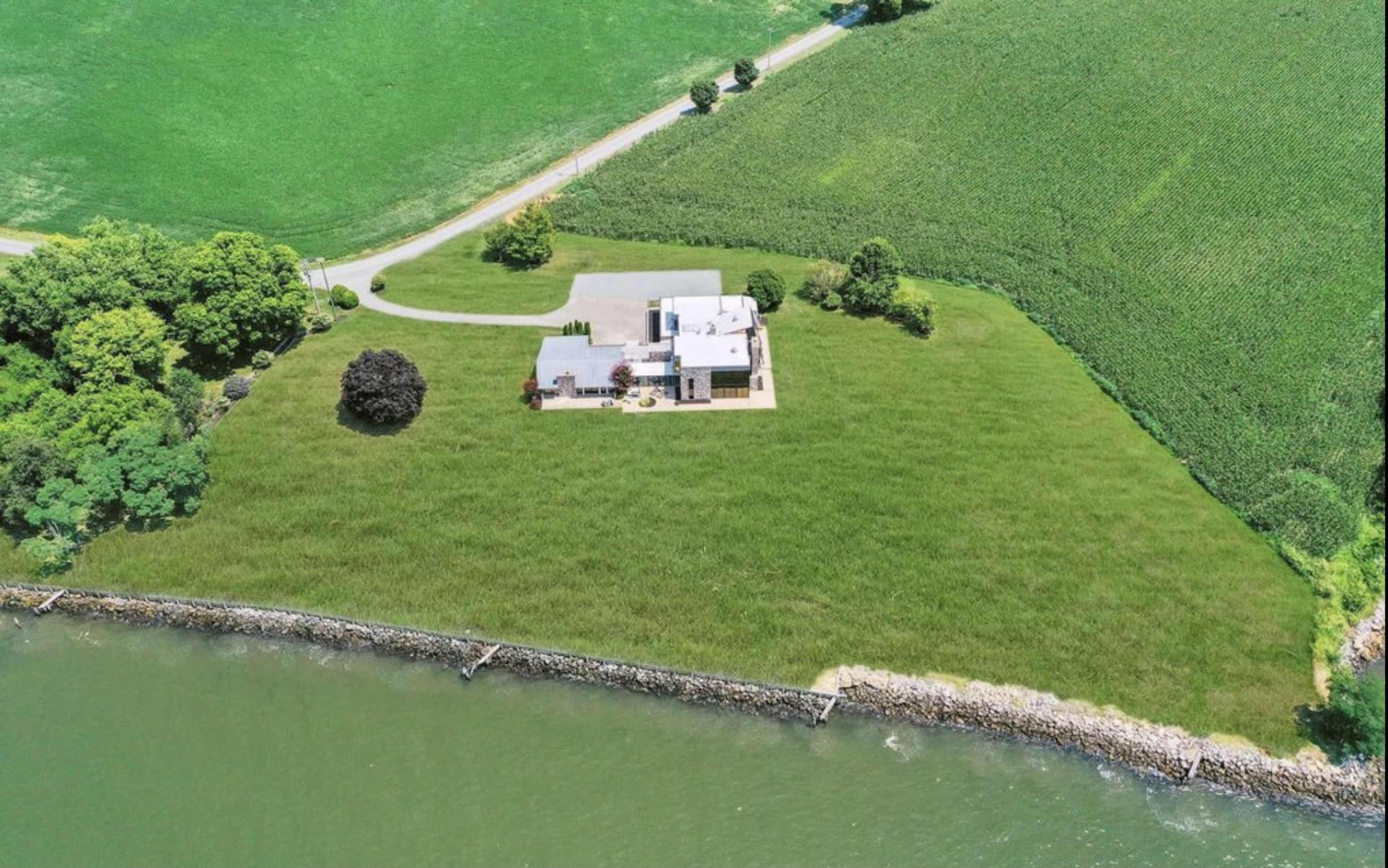 A house is situated on a large grassy area beside a body of water, with a road visible in the background and fields of crops extending beyond the property.
