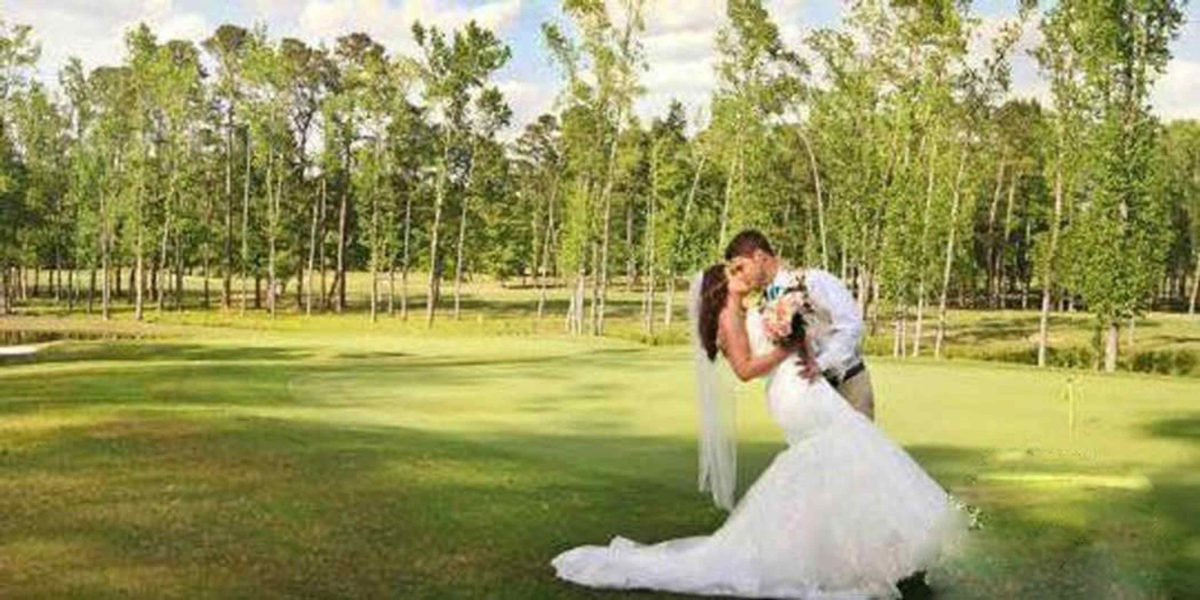 A newlywed couple embraces on a grassy area surrounded by trees.
