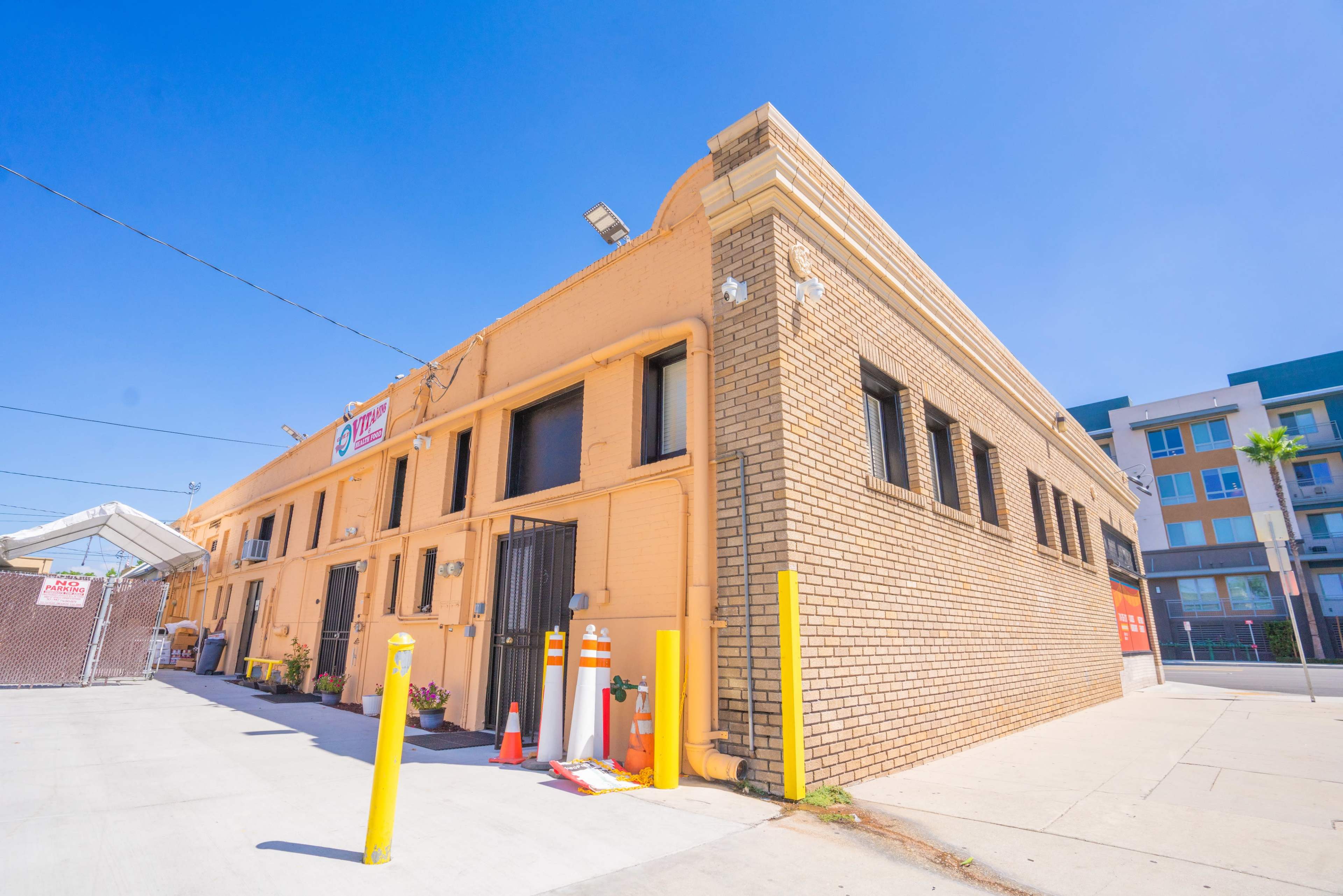 A yellow and brown brick building with a sign above the entrance is situated on a sidewalk, flanked by traffic cones and a chain-link fence.