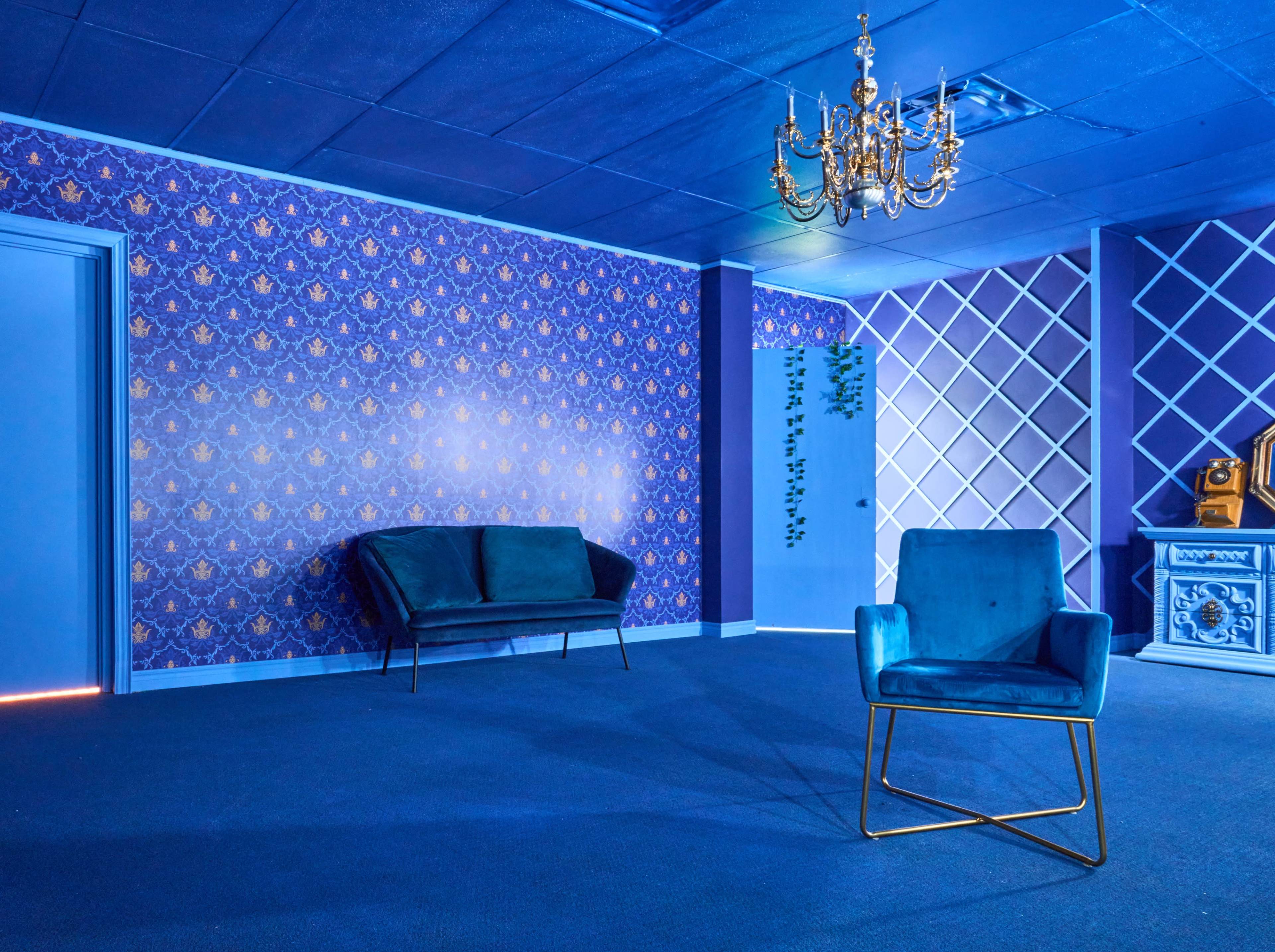 The room features a blue color scheme with patterned wallpaper, a chandelier, and a mix of seating options, including a velvet sofa and a modern chair.