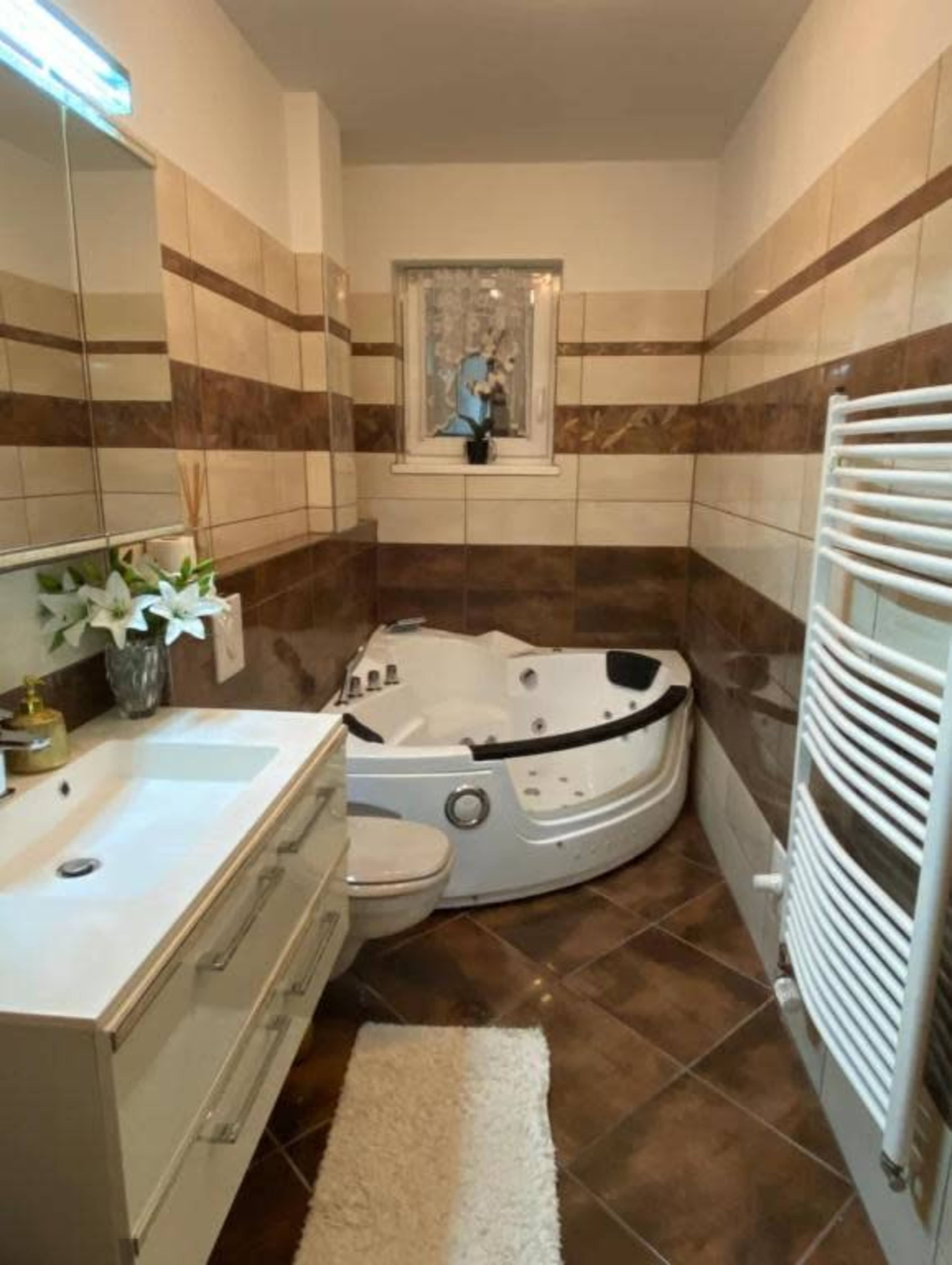 The image shows a modern bathroom with a corner jacuzzi, a vanity with a mirror, and a towel radiator on the wall.